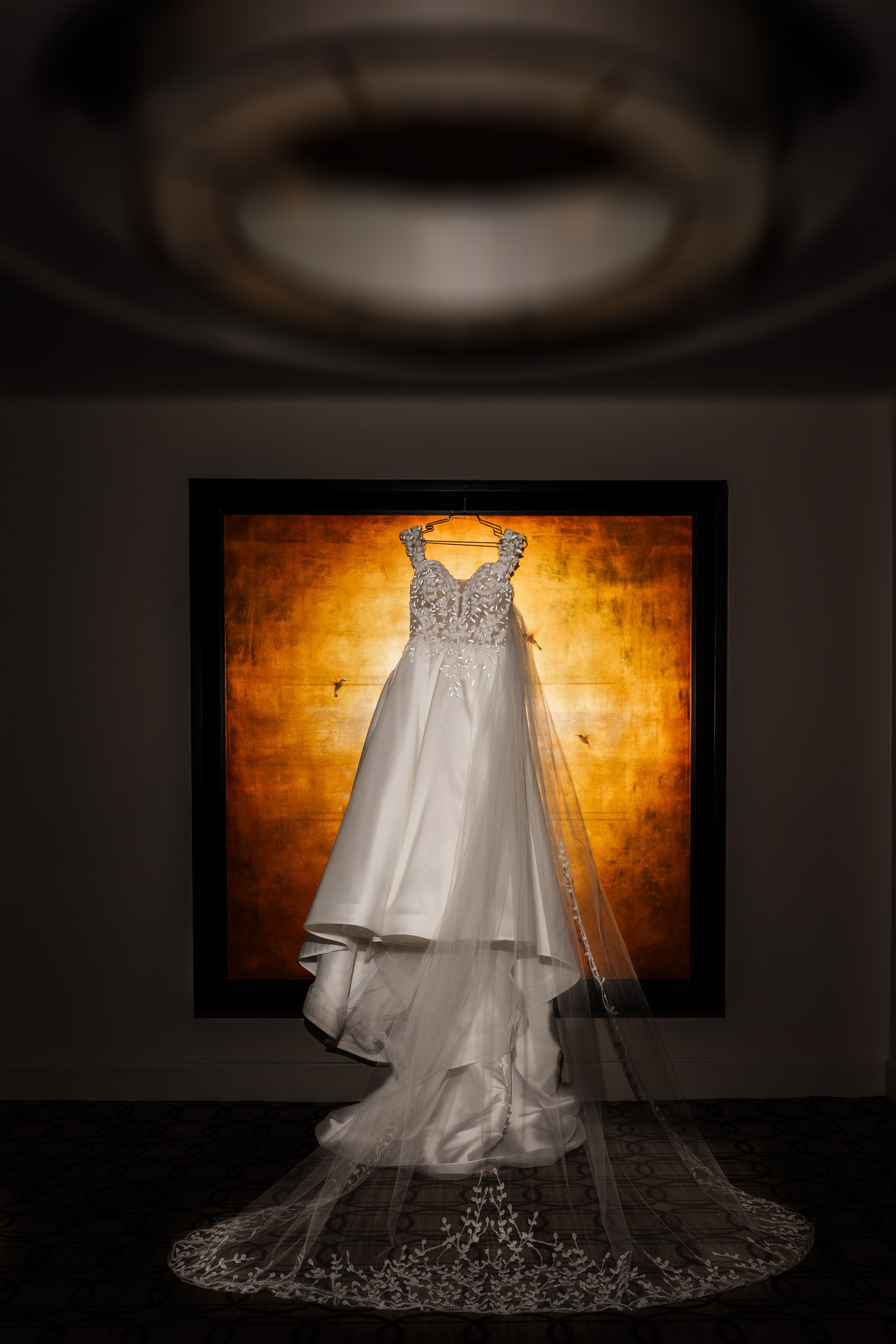 A bridal wedding gown hanging in front of a warm, glowing background. The dress has lace bodice and a long train with embroidered veil.
