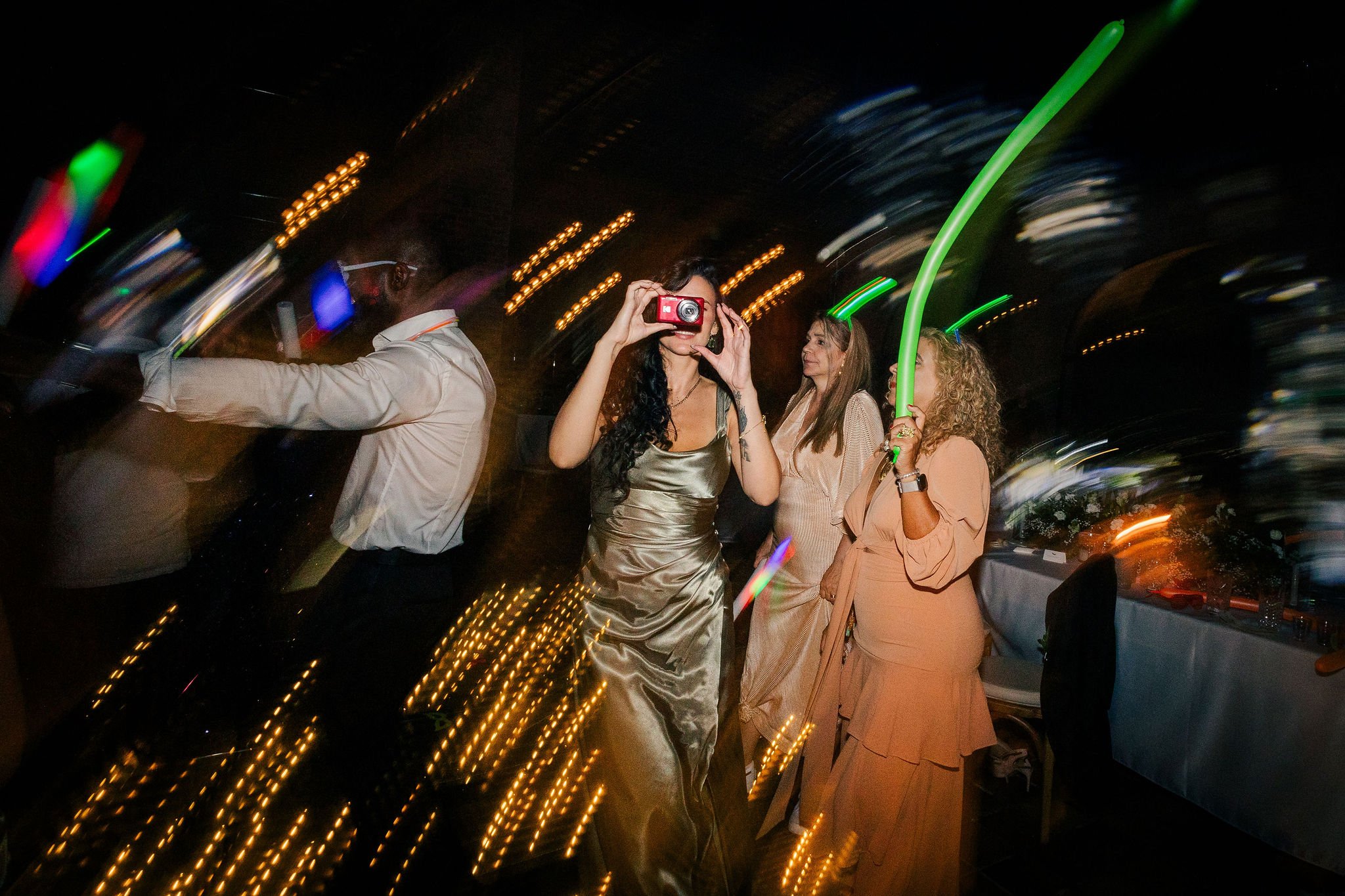 Group of women and a man dancing at a party with colorful lights, with one woman taking a photo and another holding a green light stick.