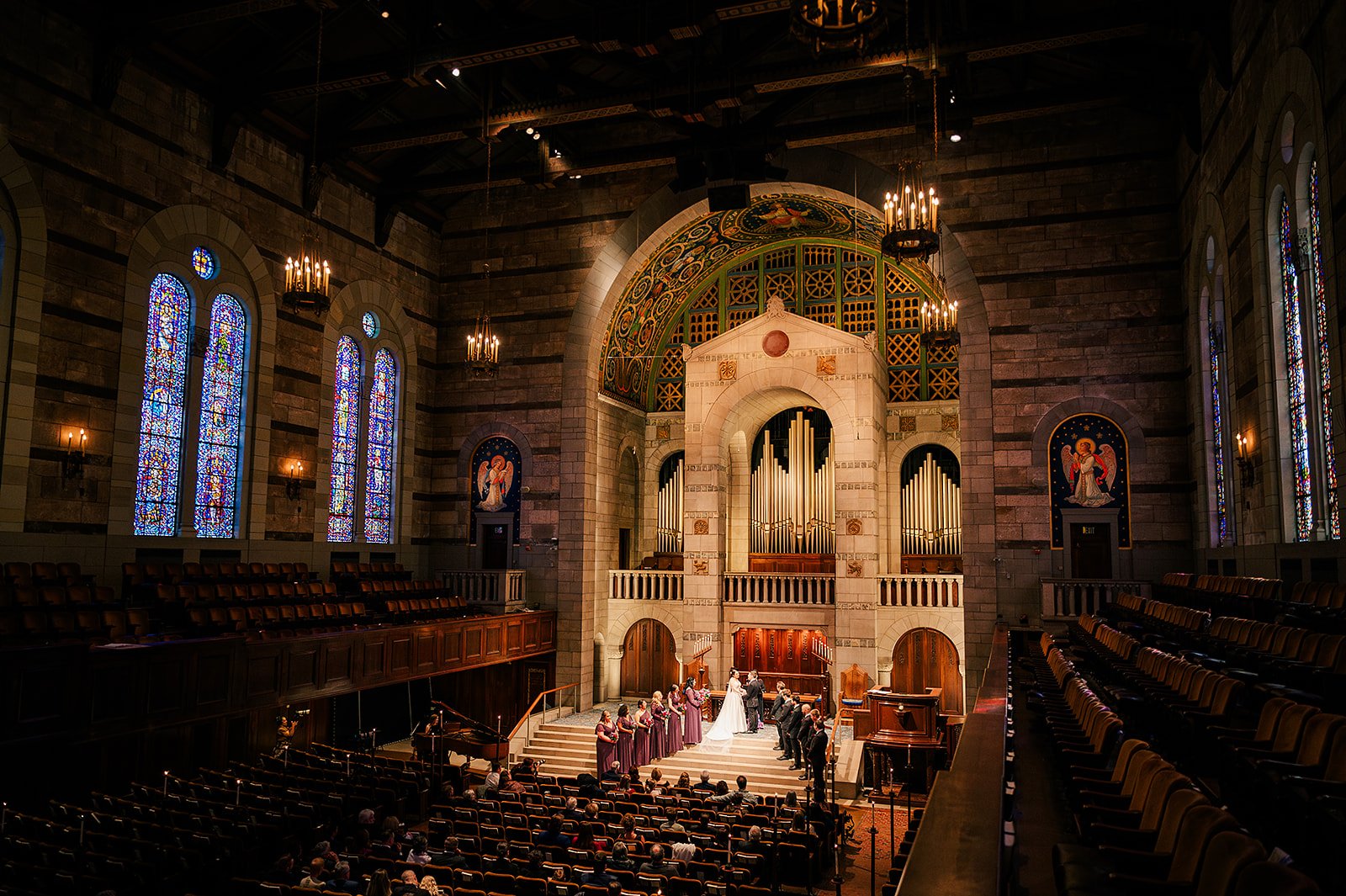 A wedding ceremony taking place in a church with stained glass windows, a pipe organ, and a congregation seated in wooden pews.