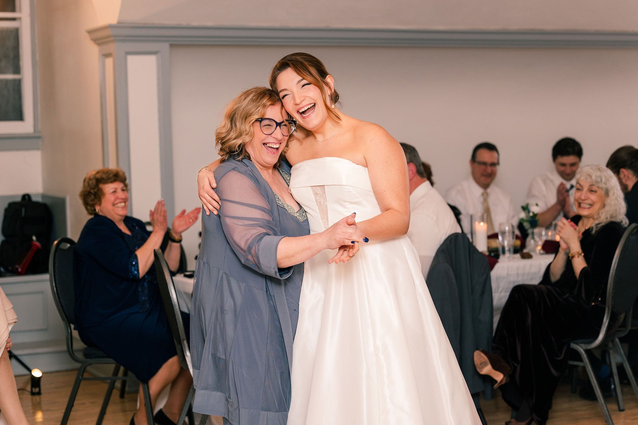 A bride in a white wedding dress dancing and hugging an older woman in a gray dress at a wedding reception, with guests sitting at decorated tables in the background, clapping and smiling.