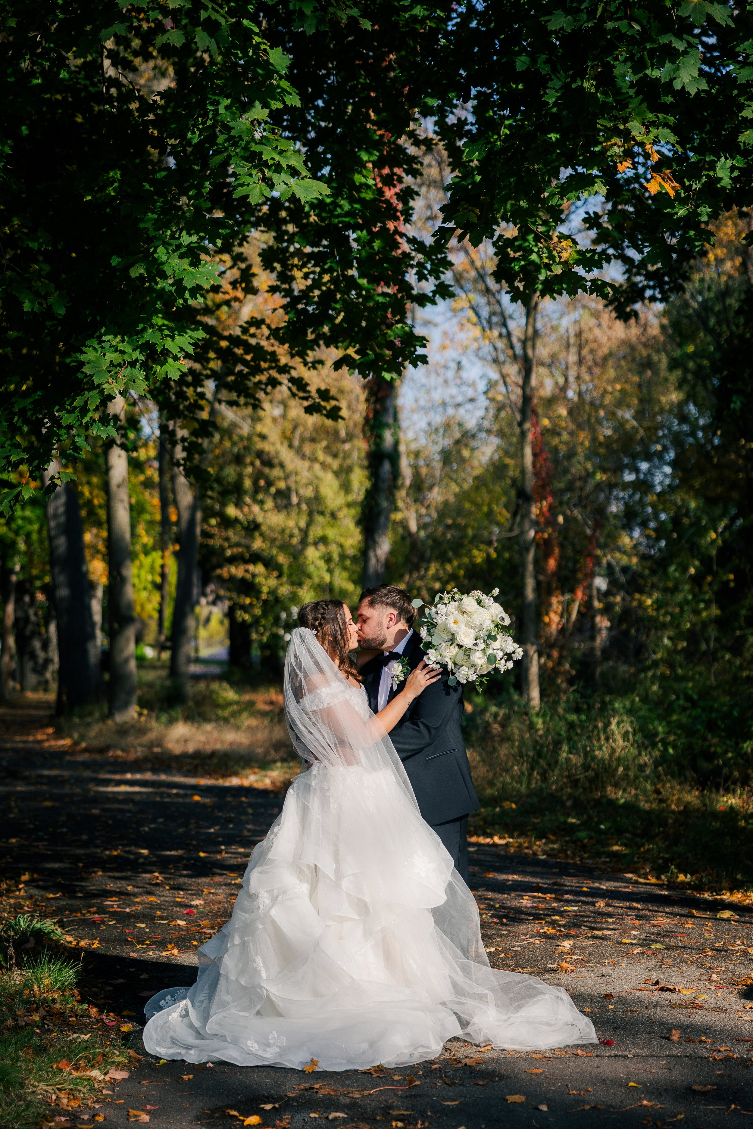 A bride and groom sharing a kiss on a wooded path, with the bride wearing a white wedding gown and veil, and the groom in a dark suit, holding a bouquet of white flowers.