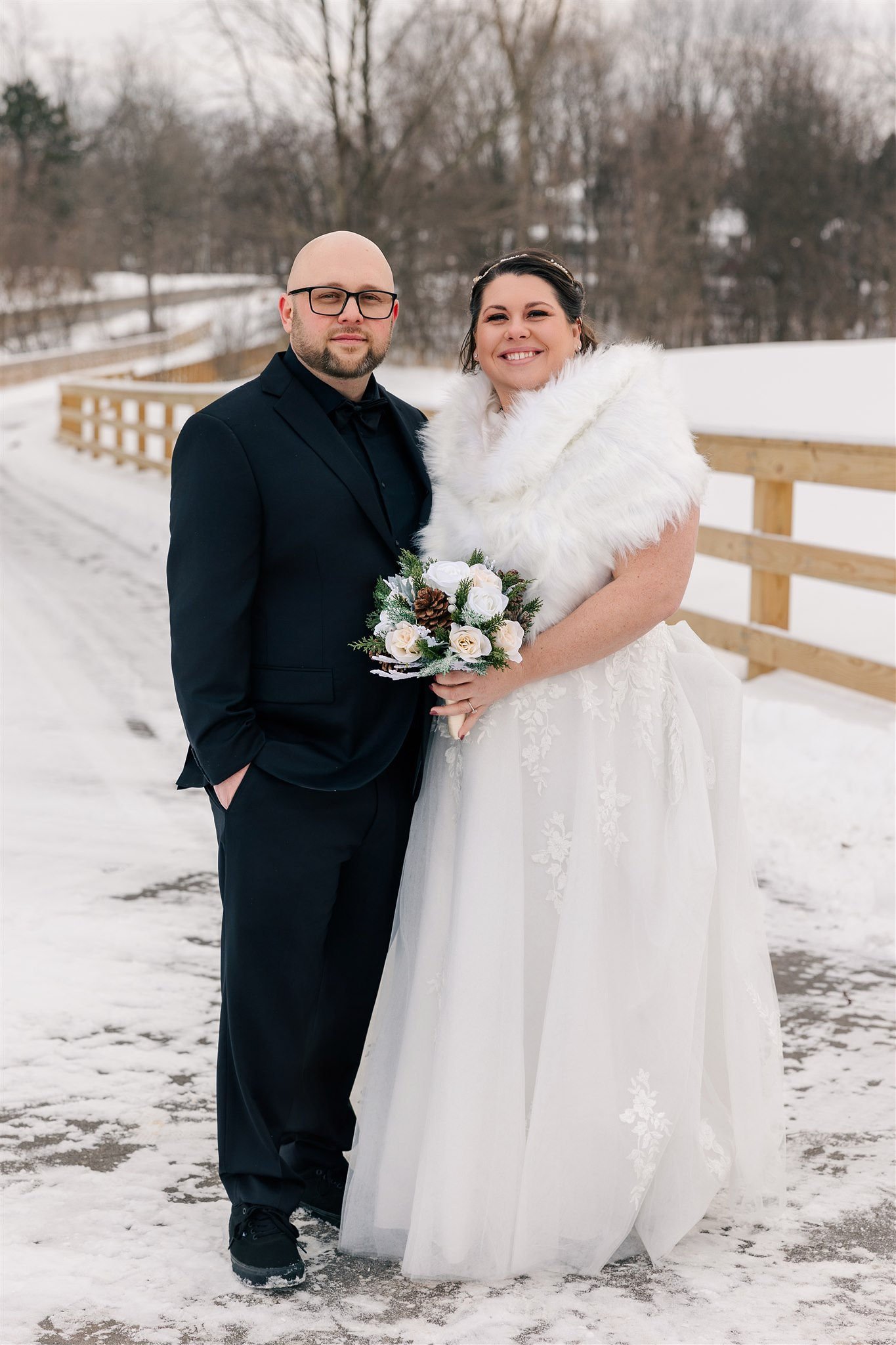 A bride and groom standing outdoors in winter, dressed in wedding attire, with the bride holding a bouquet of white roses and pinecones.