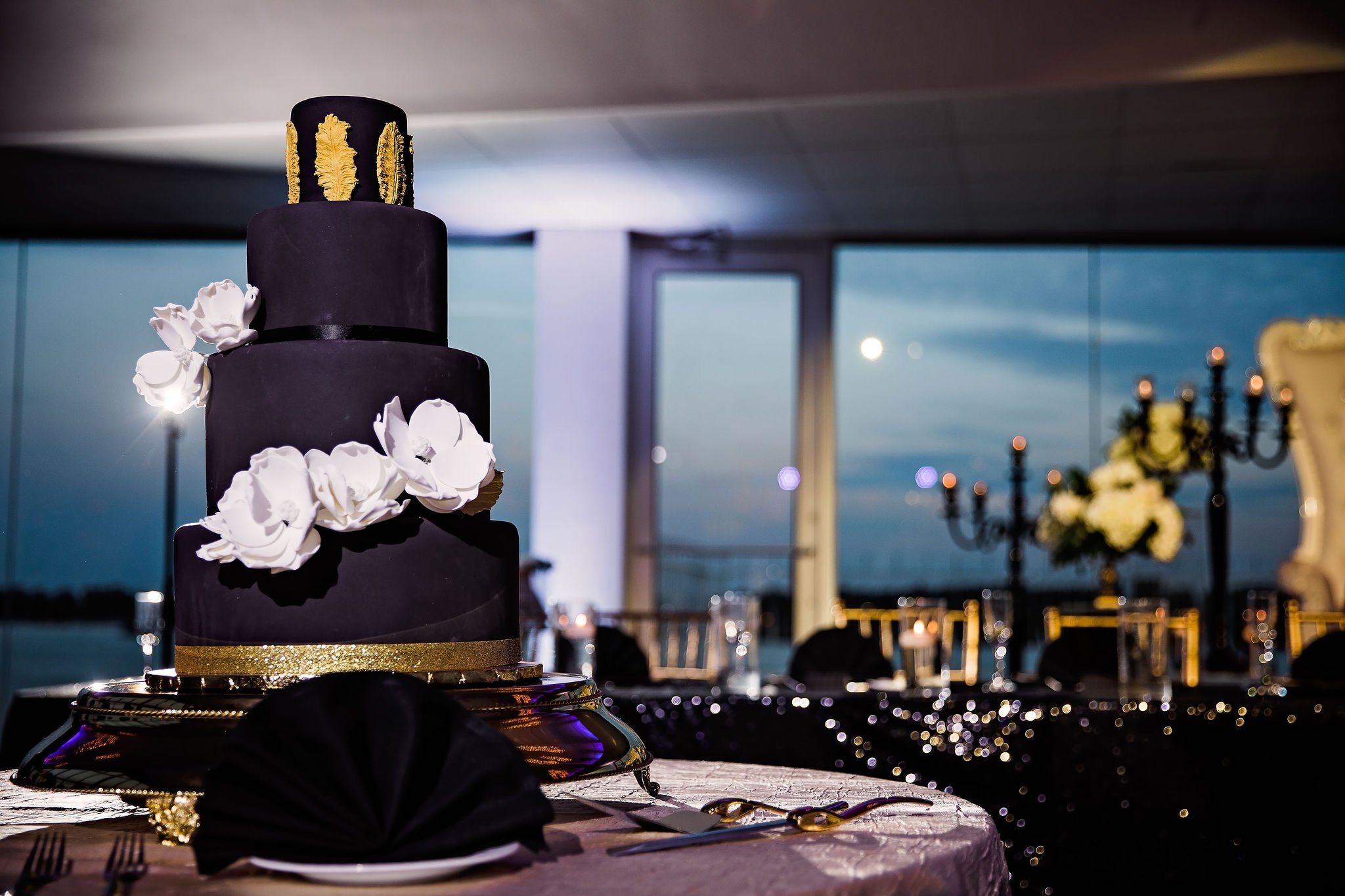 Elegant black wedding cake with white flowers and gold accents, set on a table with black napkins and gold utensils, overlooking a waterfront at dusk with chandeliers and floral arrangements in the background.