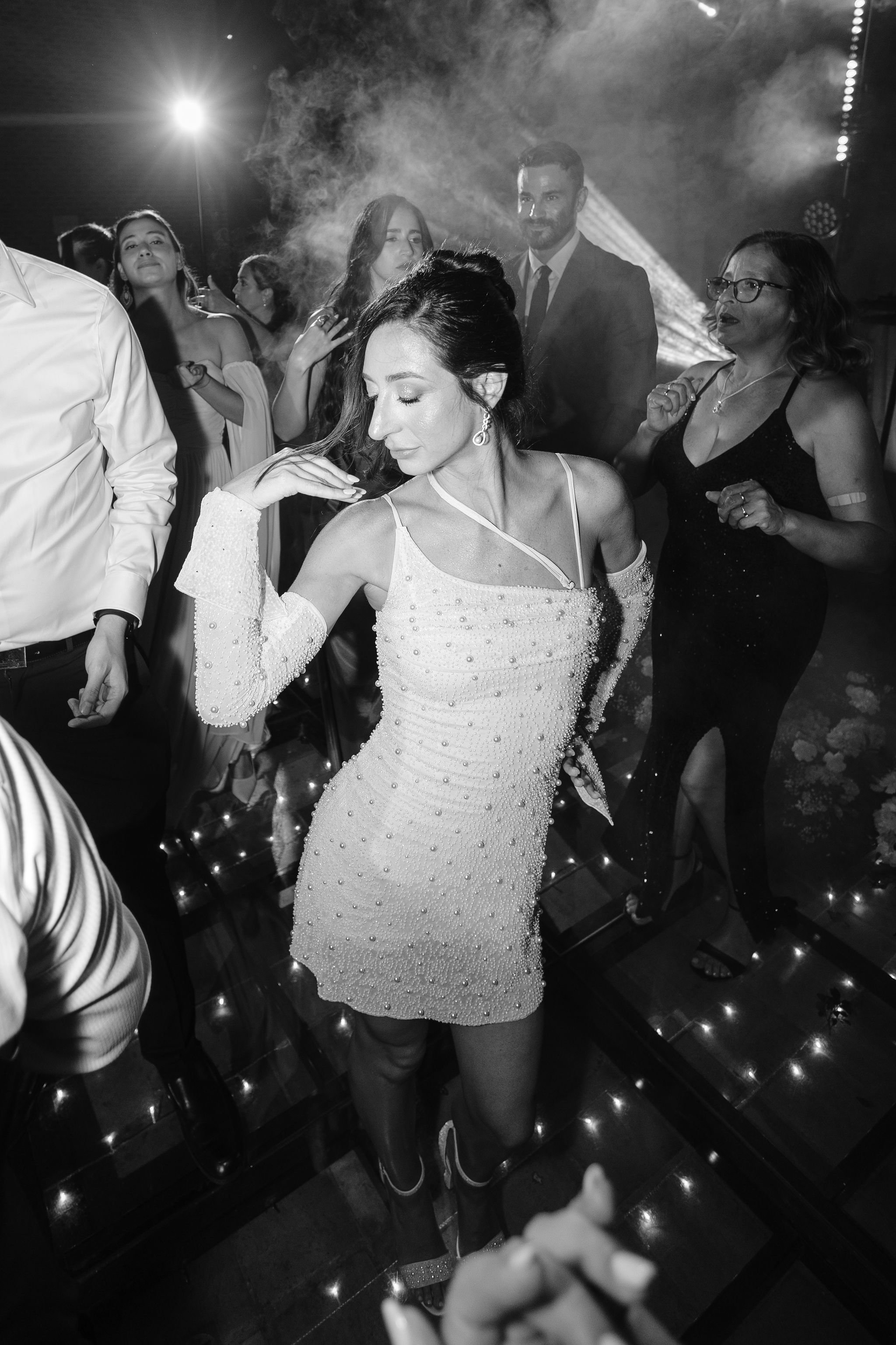Woman in a white, pearl-studded dress dancing at a party or nightclub, surrounded by other guests, with dramatic lighting and smoke effects in the background.
