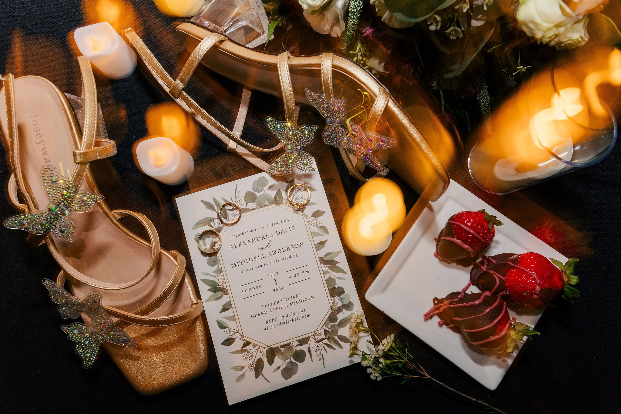 Golden high heel shoes with butterfly embellishments, wedding invitation, gold rings, candles, strawberries dipped in chocolate, and flowers on a black surface, celebrating a wedding.