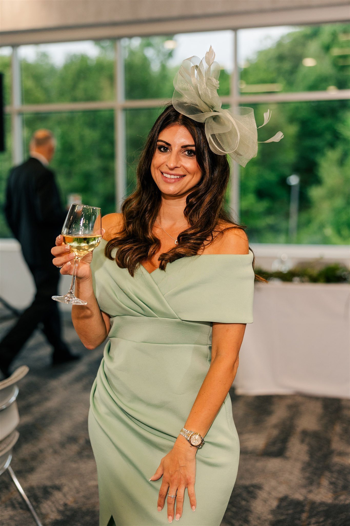 A woman in a light green off-shoulder dress and large fascinator hat holding a glass of white wine at an indoor event with large windows showing green trees outside.