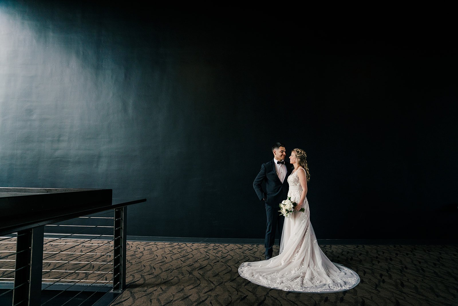 Bride and groom standing together in a minimalist setting, with the bride holding a bouquet, both dressed in wedding attire, facing each other, against a dark, empty background.