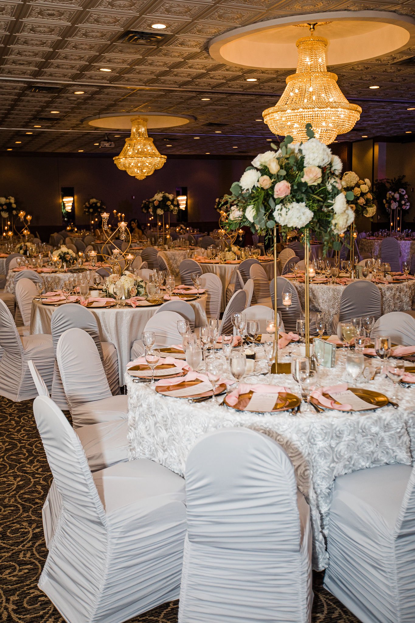Elegant banquet hall set up for a wedding reception with round tables covered in white tablecloths, adorned with pink napkins, floral centerpieces, and tall candle holders, illuminated by large chandeliers.