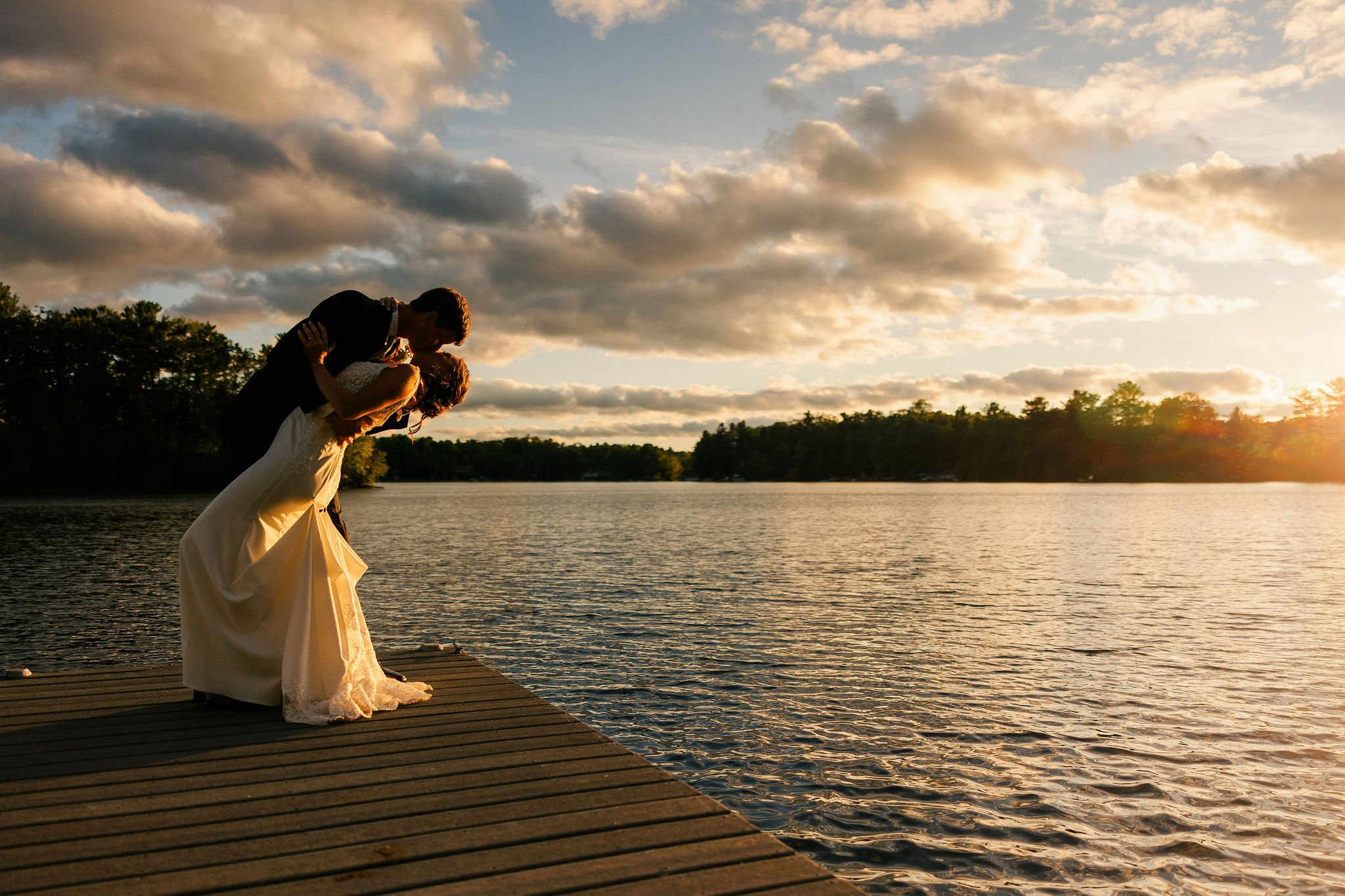 A bride and groom sharing a kiss at sunset on a dock by a lake.