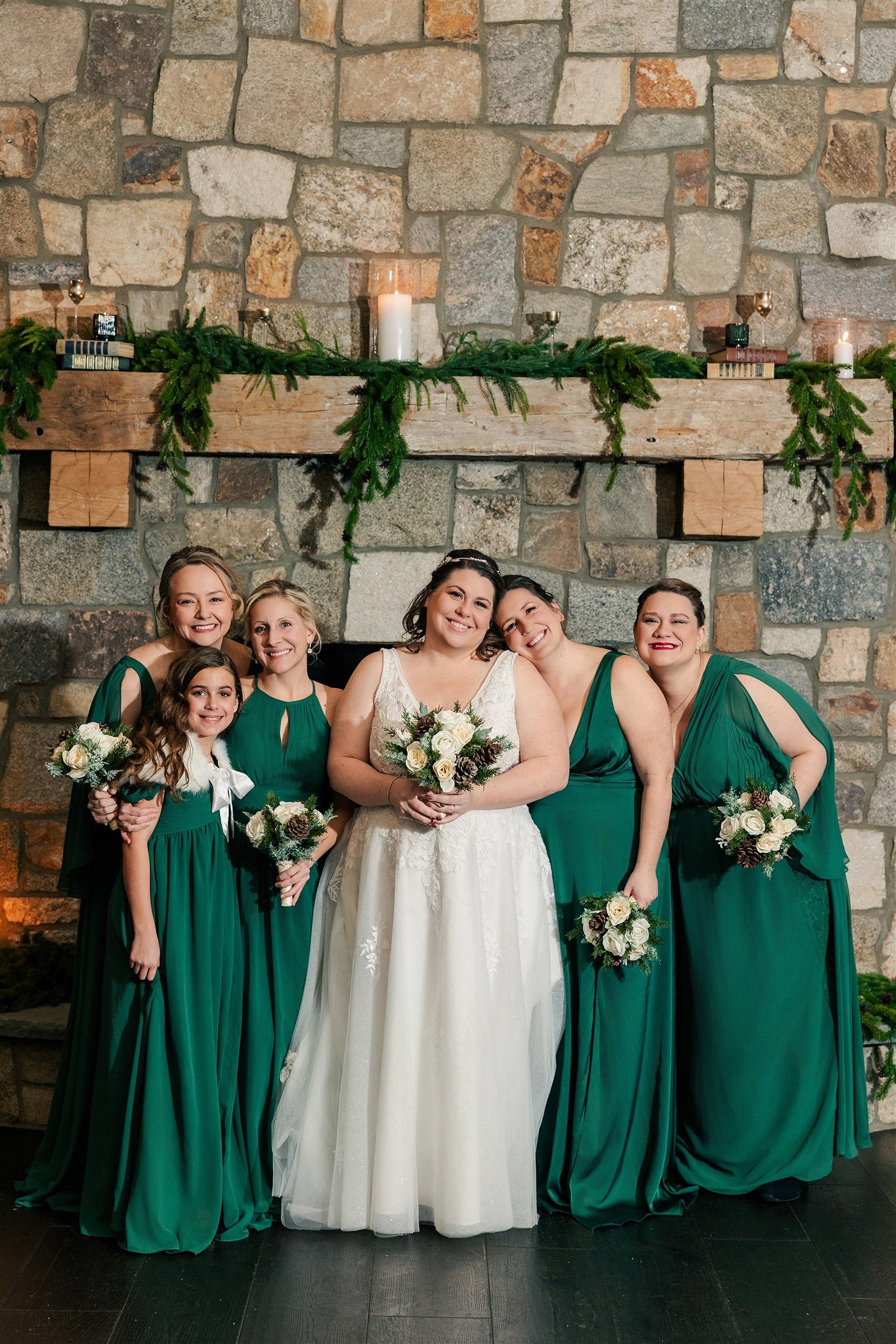 A group of women, including a bride in a white wedding dress, and four bridesmaids in matching green dresses, standing in front of a stone fireplace decorated with greenery, candles, and books at a wedding celebration.