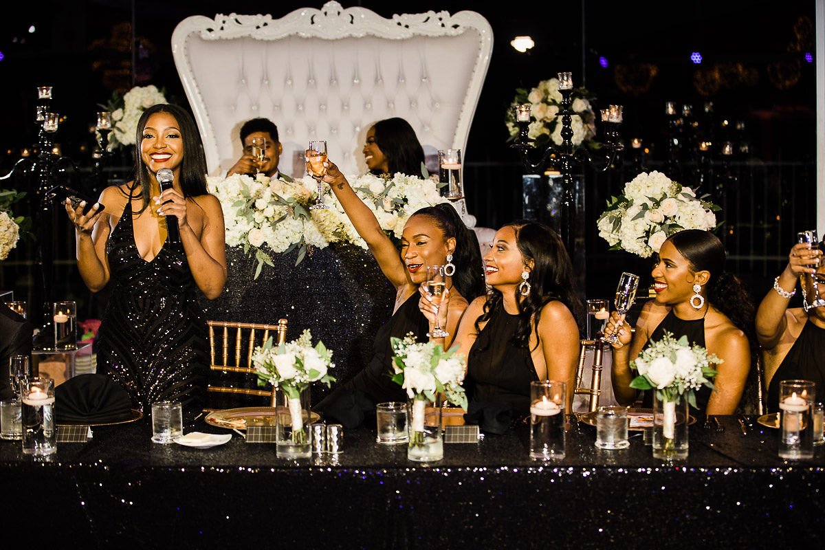A woman in a black dress speaking into a microphone while a group of women in black dresses seated at a decorated table raise glasses in a toast at an elegant event or celebration.