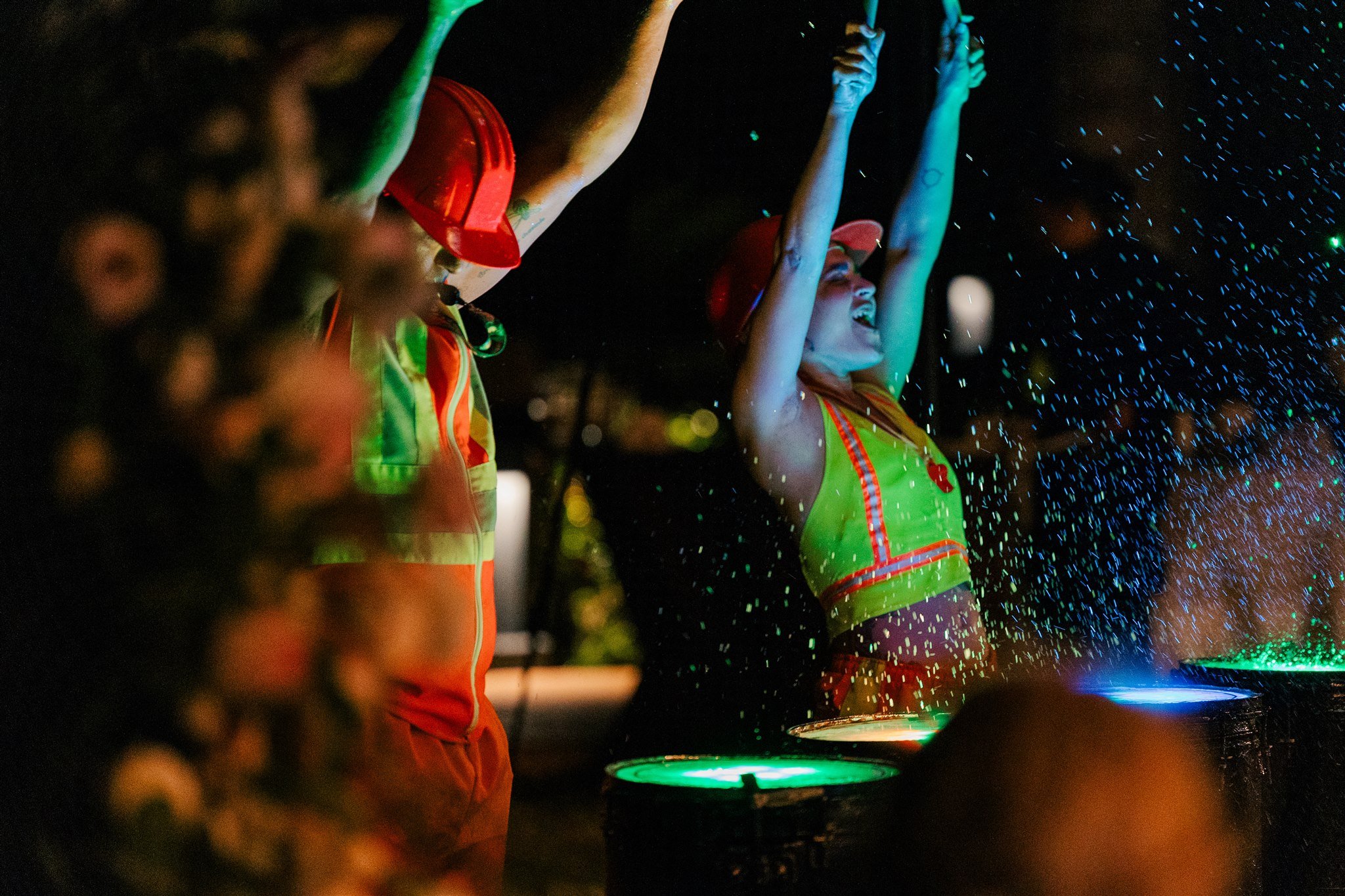 People celebrating at night, spraying colorful water or paint from drums or buckets, wearing bright vests and red hats.