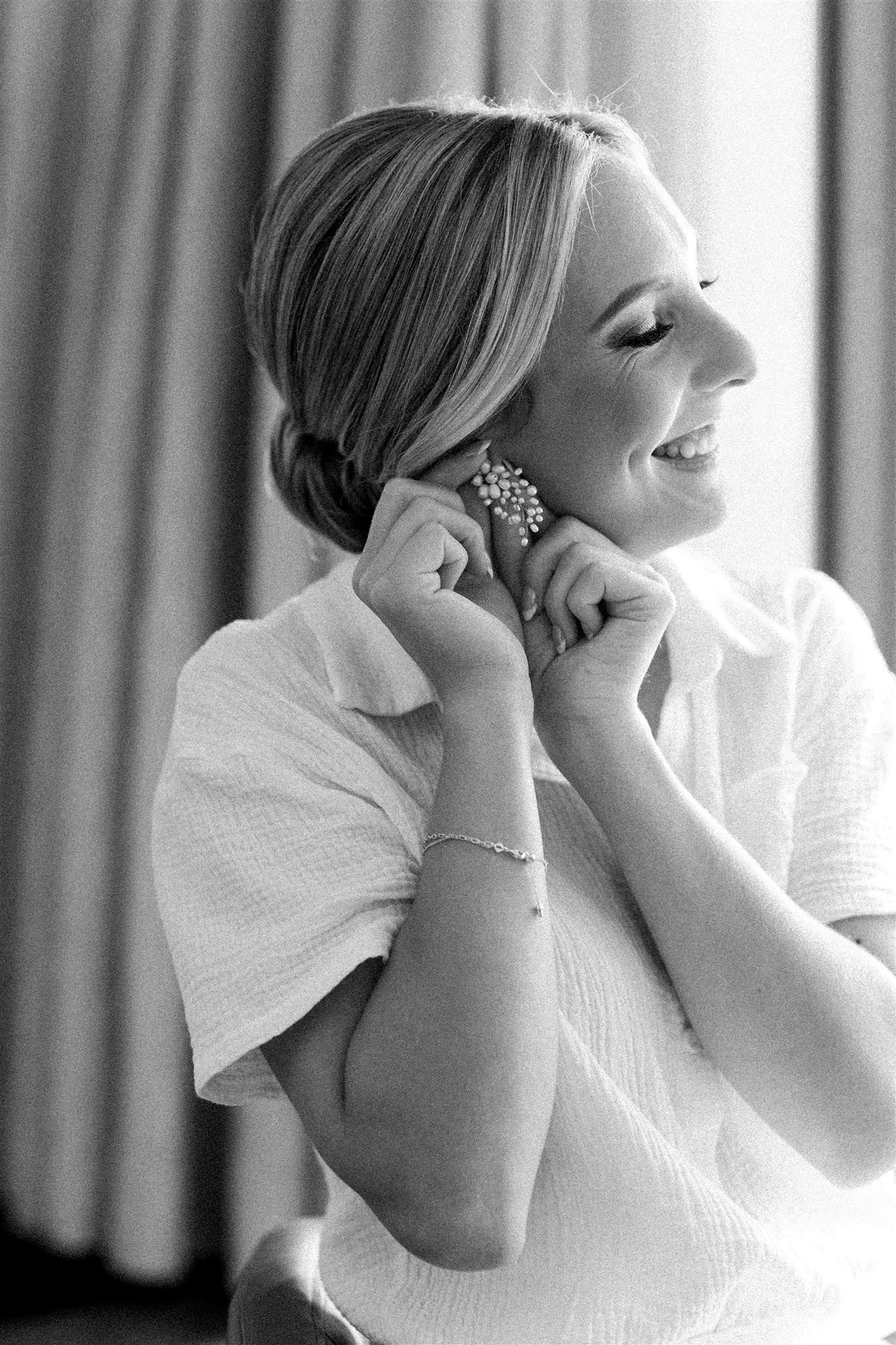 A smiling woman with short hair, dressing in a light-colored shirt, putting on a large, ornate earring, with a curtain in the background.