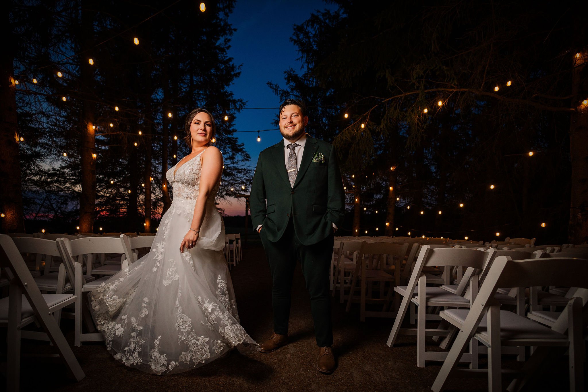 Bride in a lace wedding gown and groom in a green suit with a tie standing outdoors at dusk, illuminated by string lights and surrounded by empty white chairs under tall trees.