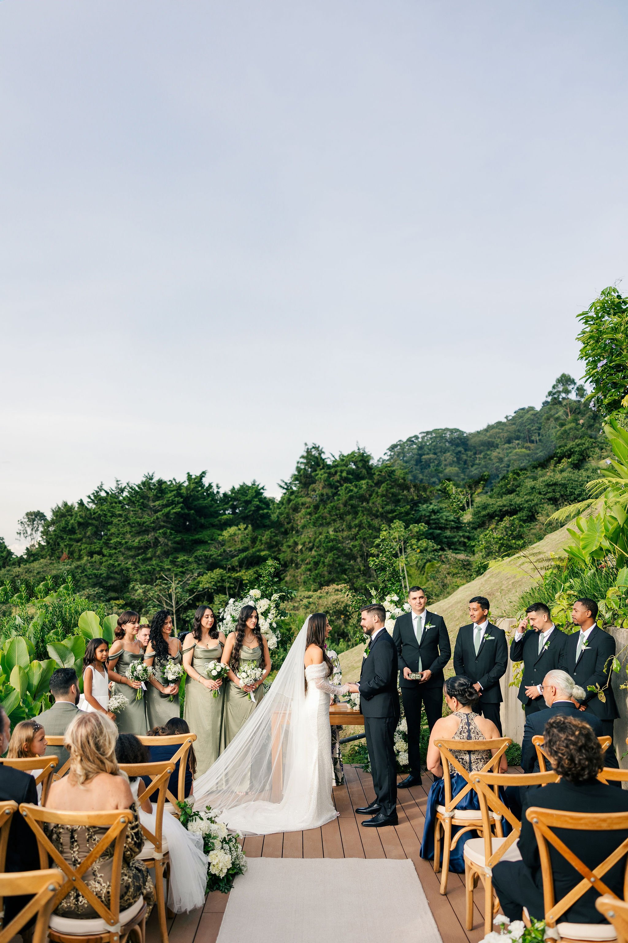 Bride and groom exchanging vows during an outdoor wedding ceremony on a wooden deck surrounded by lush green trees and hills, with bridesmaids and groomsmen standing nearby, and guests seated in wooden chairs watching.
