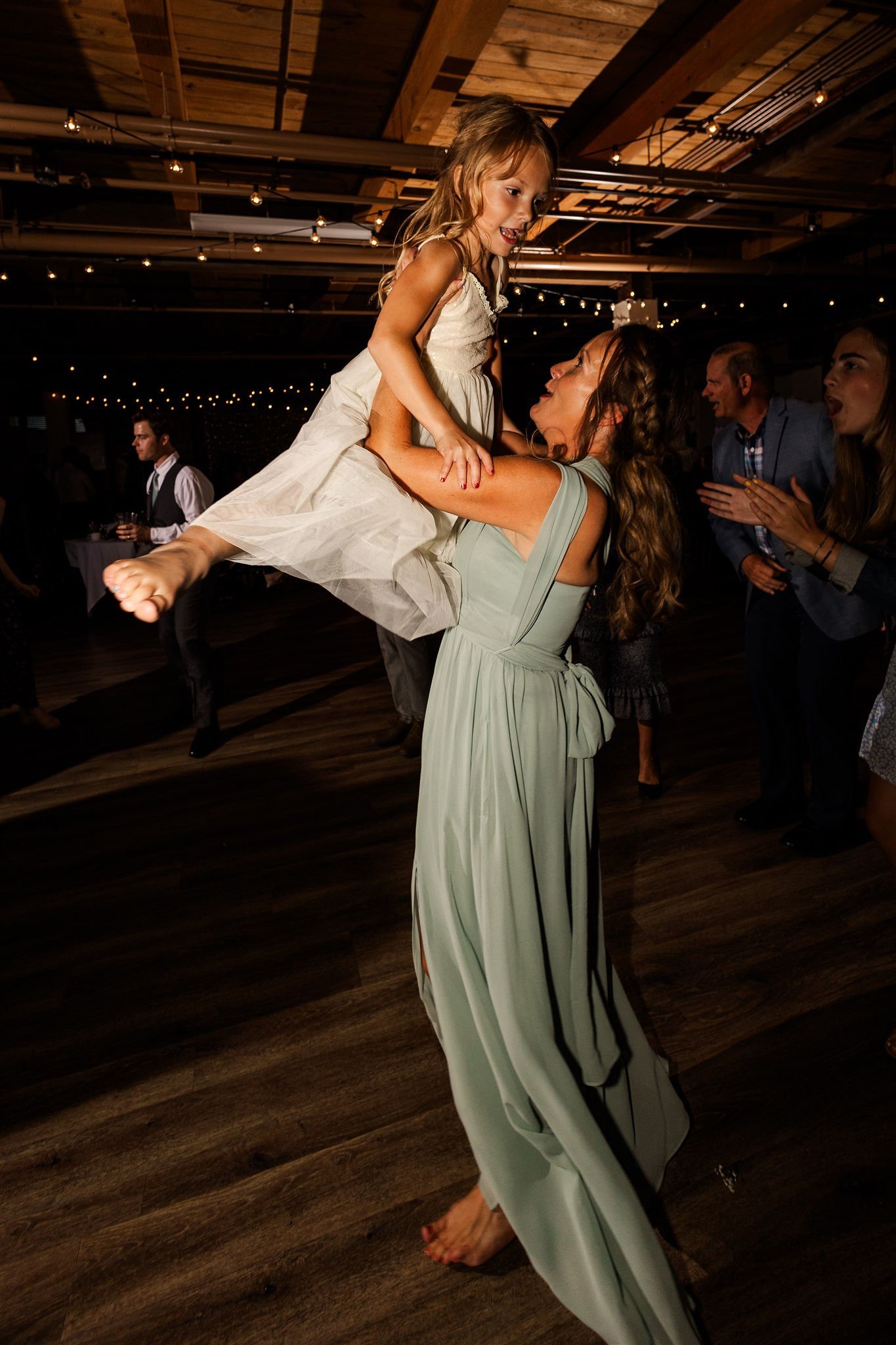 A woman dressed in a light green dress lifting a young girl in a white dress during a celebration or dance party, with other guests in the background under string lights.
