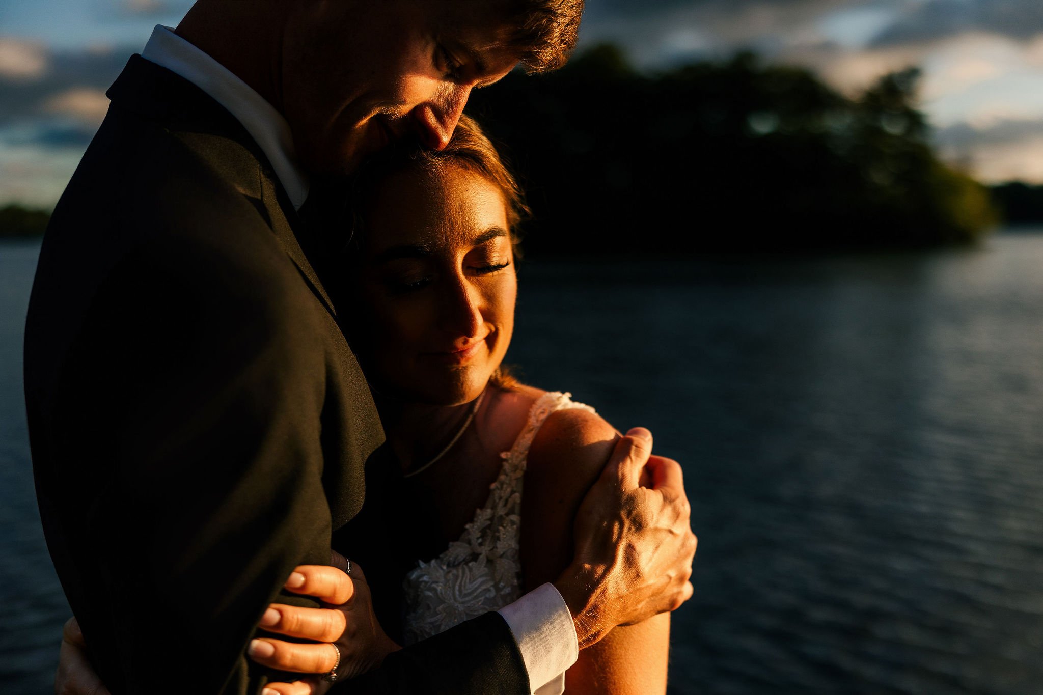 A man and woman embrace lovingly outdoors at sunset, close-up shot with water and trees in background.