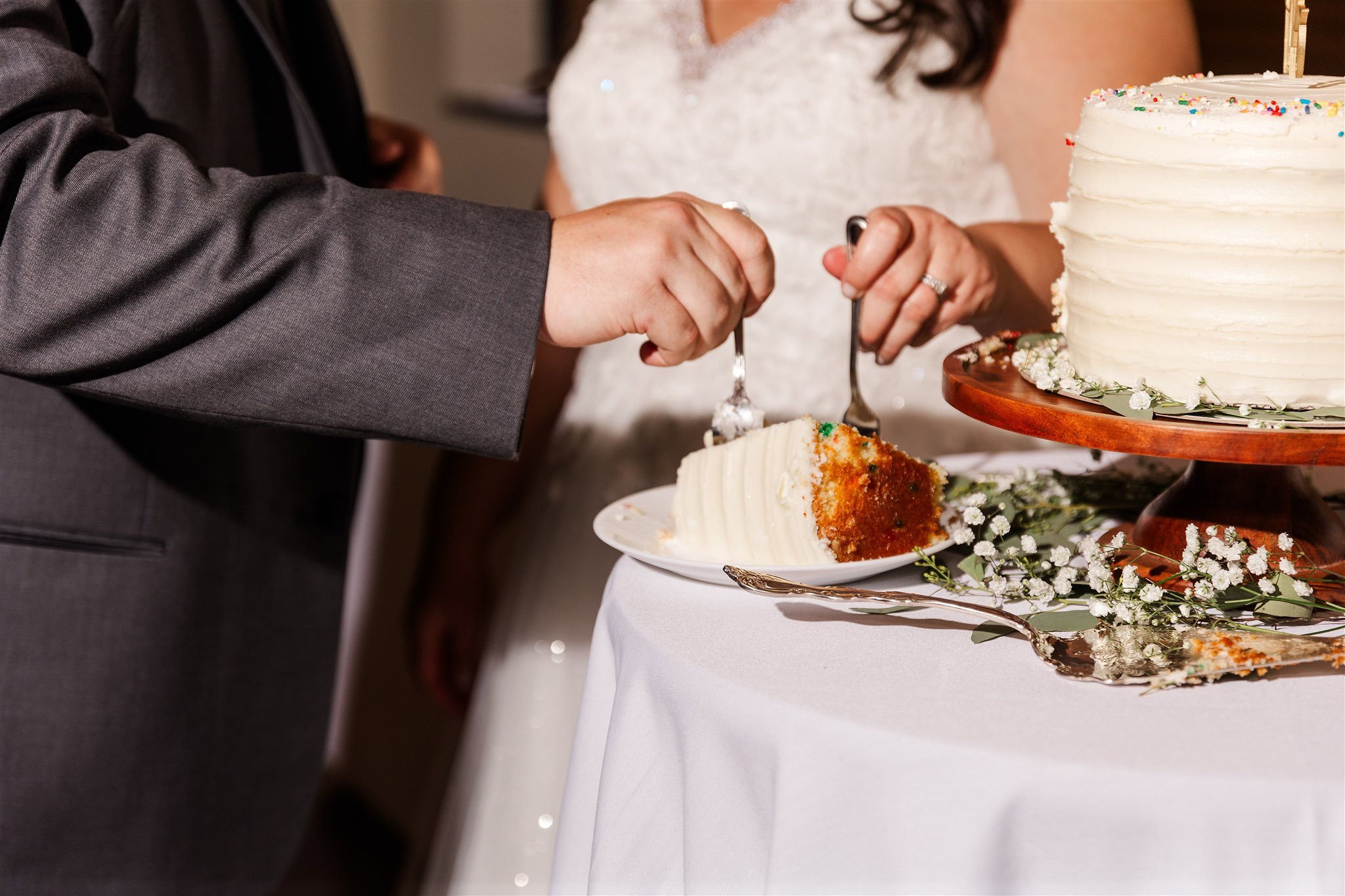A newlywed couple cuts a slice of wedding cake together at their reception. The cake is on a table with floral decorations.