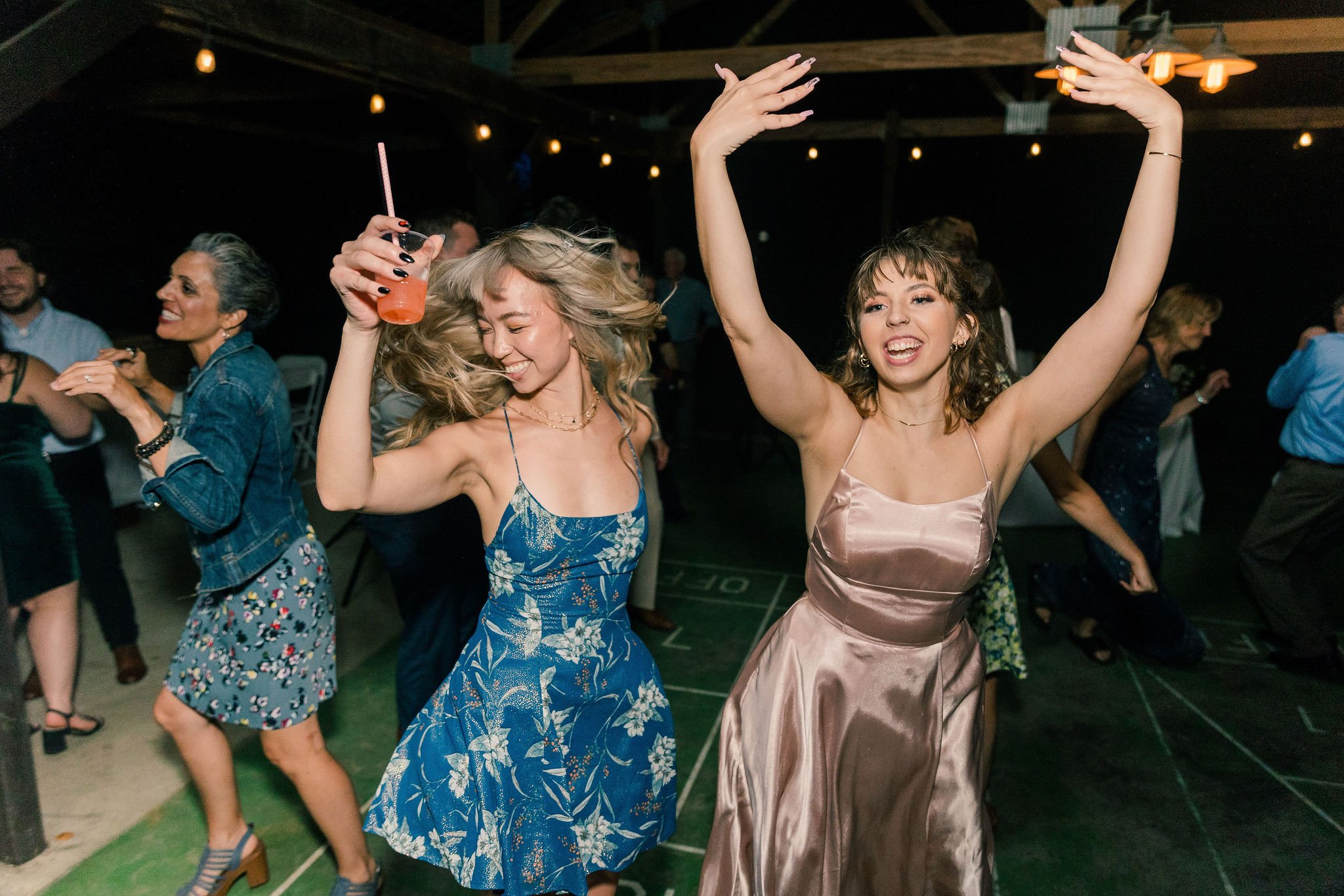 People dancing and celebrating at a party or wedding reception, with two young women in the foreground joyfully dancing and smiling, wearing elegant dresses, one holding a drink.