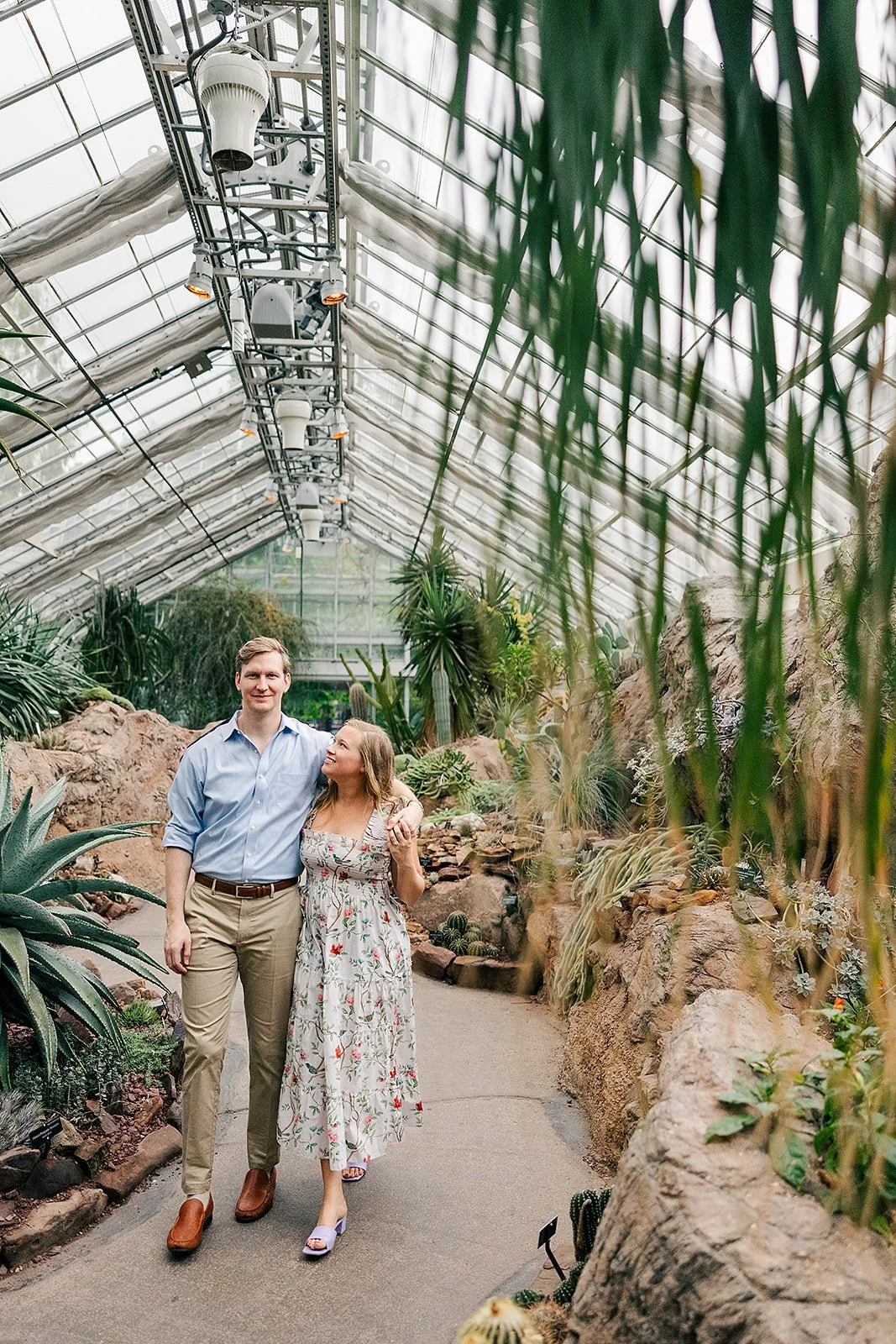 A couple walking through a lush greenhouse filled with various plants and cacti, with a glass ceiling overhead.