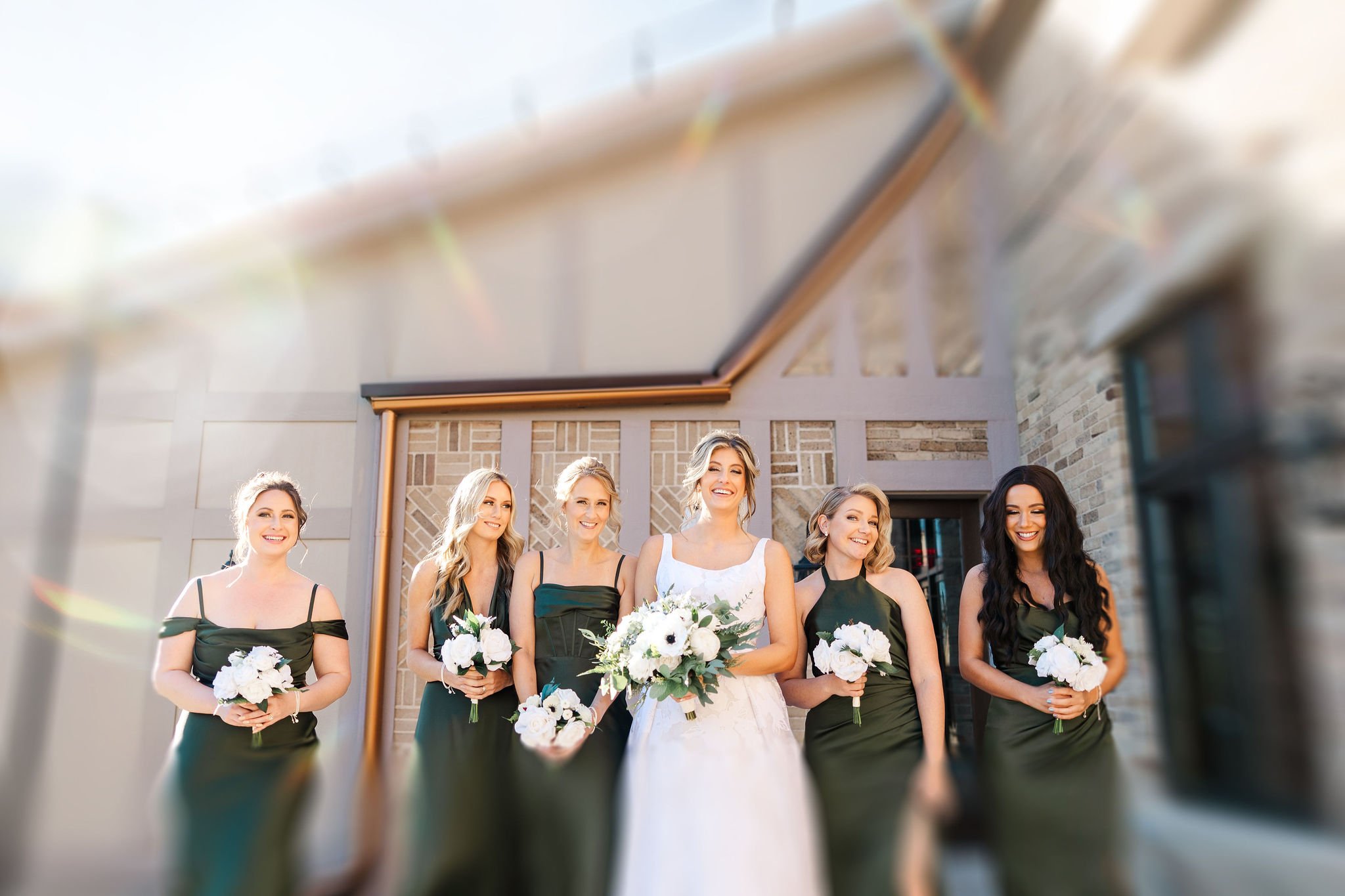 Bridal party with the bride and five bridesmaids holding bouquets, standing outside in front of a building with brick and wood siding, sunny weather.