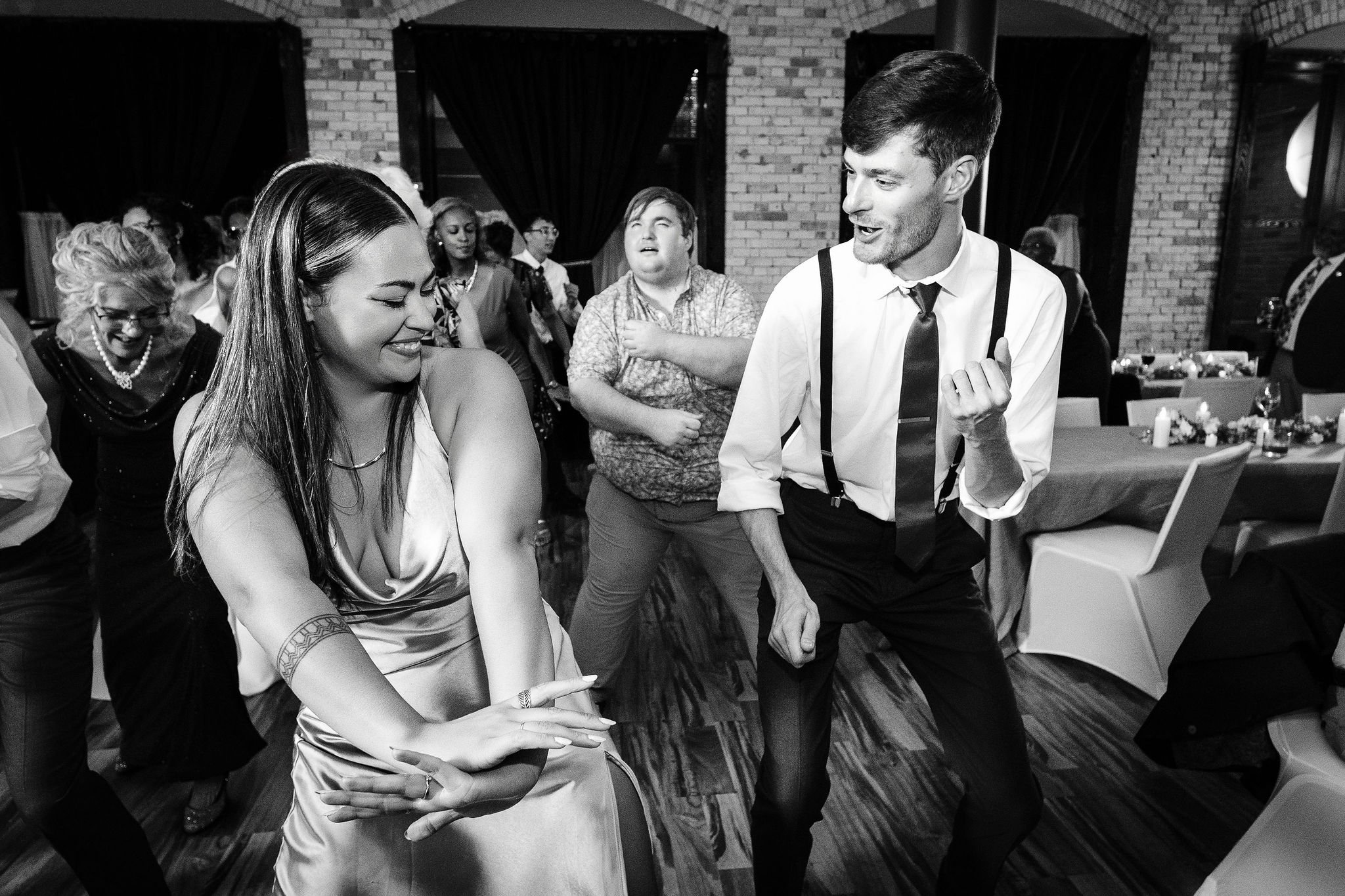 People dancing at a celebration or party, smiling and enjoying themselves in a decorated venue with brick walls and tables.