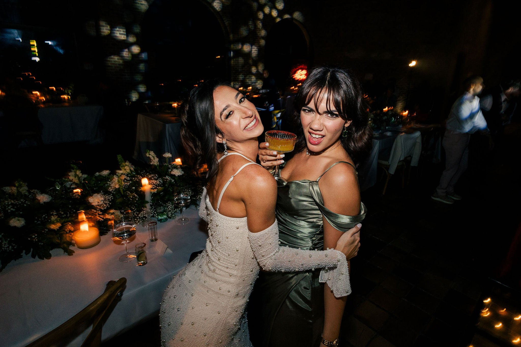 Two women at a celebration, smiling and posing with a cocktail in a dimly lit room with candles and floral decorations on the table.