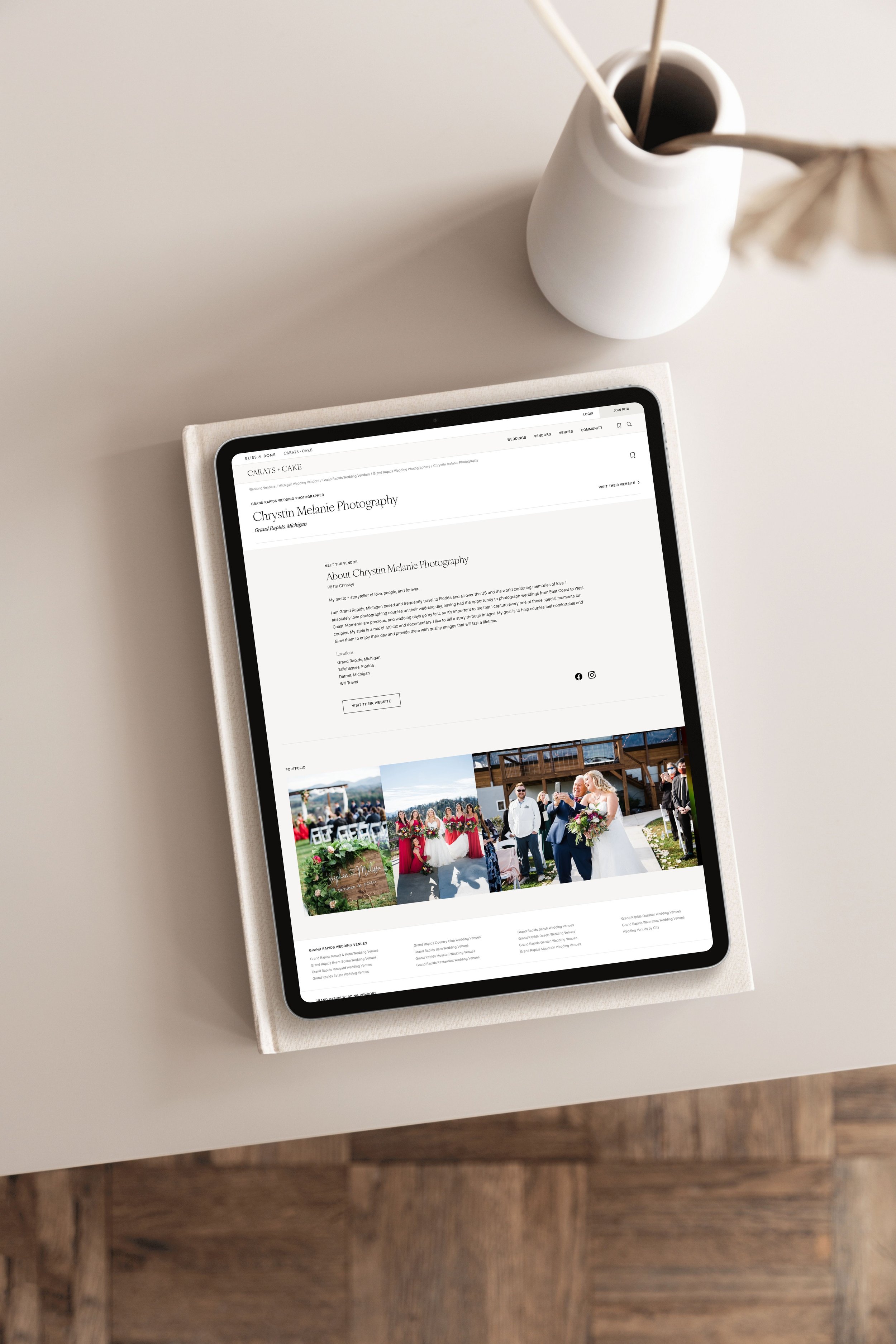 An iPad displaying a wedding photography website on a light-colored table with a white vase holding two sticks and a paper umbrella in the background.