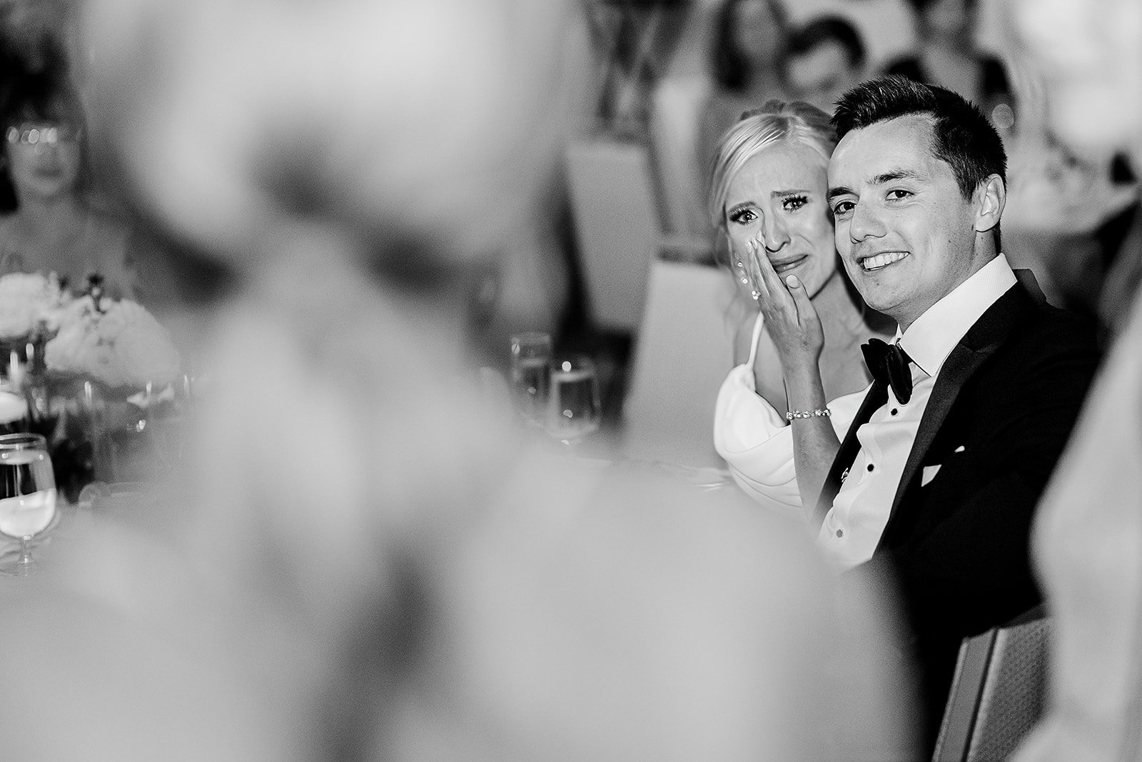 A black and white photo of a woman and a man at a formal event, with the woman appearing emotional and wiping tears from her eyes, while the man is smiling, dressed in a tuxedo, seated at a table with glasses and floral arrangements.
