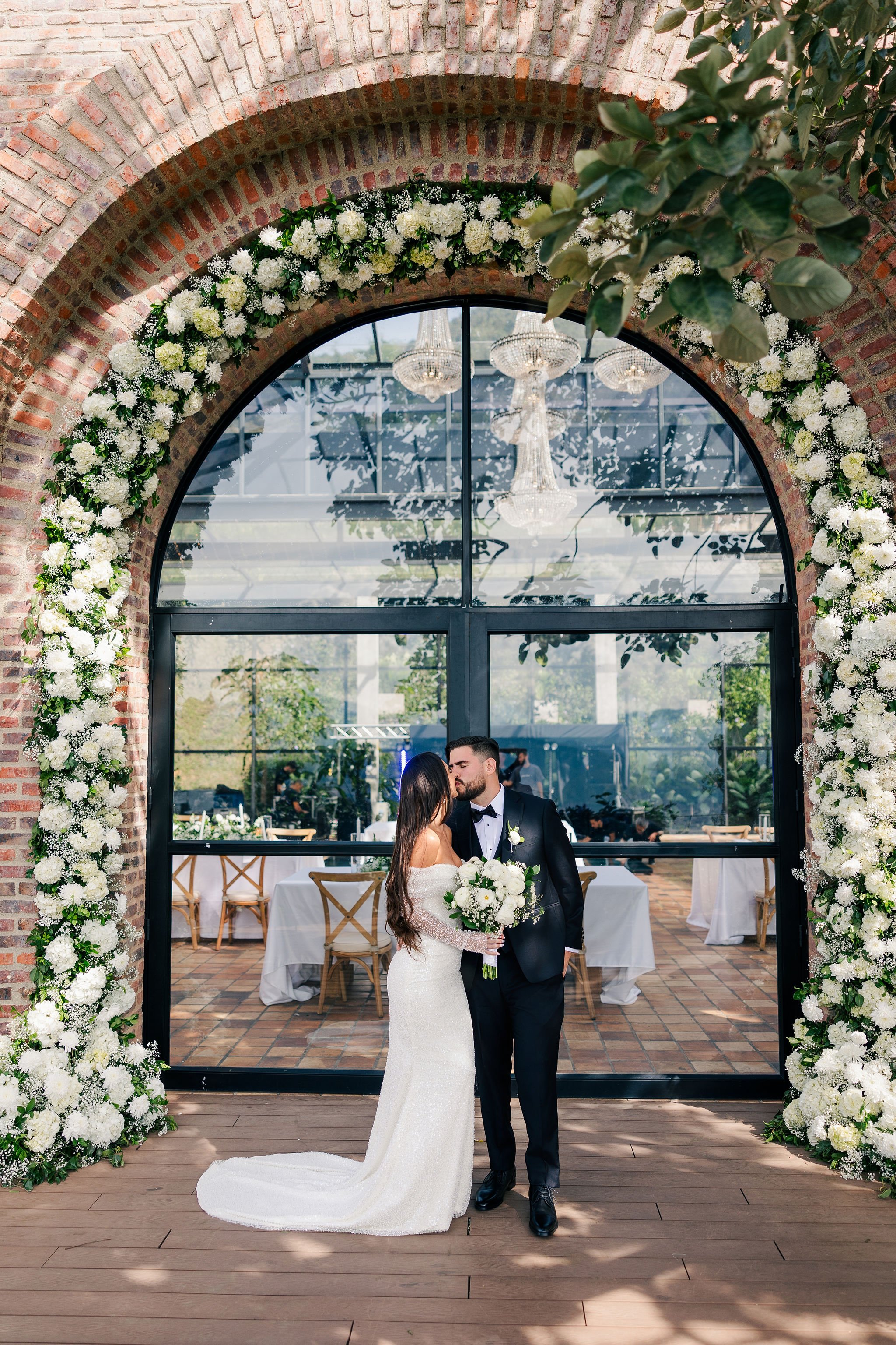 Bride and groom sharing a kiss at their wedding ceremony, standing beneath an arch decorated with white flowers. The bride is wearing a white gown, and the groom is in a black tuxedo. There is a glass window behind them with chandeliers hanging insid