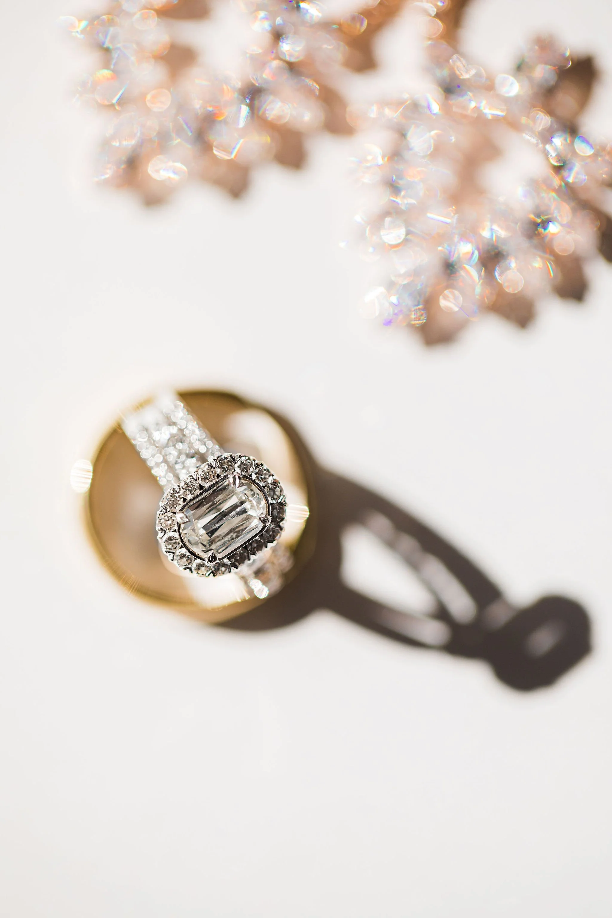 Close-up of a diamond ring with a large rectangular center diamond surrounded by smaller diamonds, casting a shadow on a white background. A blurred sparkling object is visible in the upper part of the image.