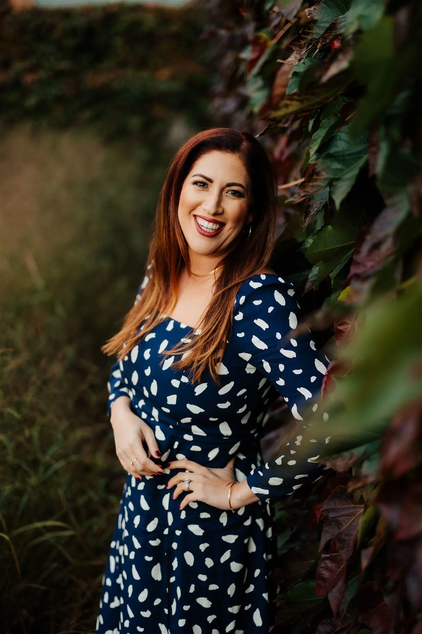 A woman with long, reddish-brown hair is smiling and wearing a navy dress with white abstract patterns. She is standing outdoors next to a wall covered in dark green and reddish leaves. The background is blurred but appears to be a natural, outdoor setting.