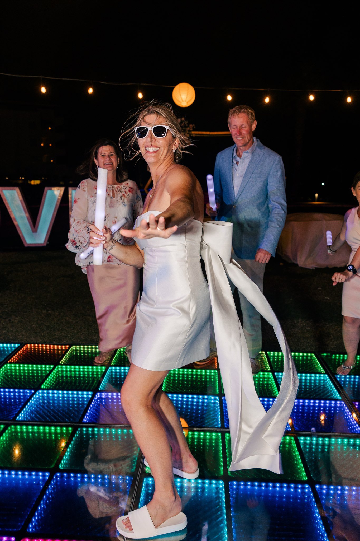 A woman in a white dress dancing on a colorful light-up dance floor at night, with other people in the background, under string lights and paper lanterns.