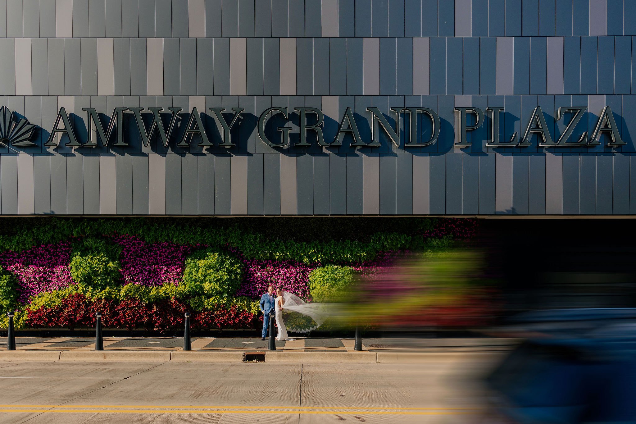A bride and groom in wedding attire standing on a sidewalk in front of an Amway Grand Plaza building with colorful flowers and green foliage behind them, and a blurred car passing by.