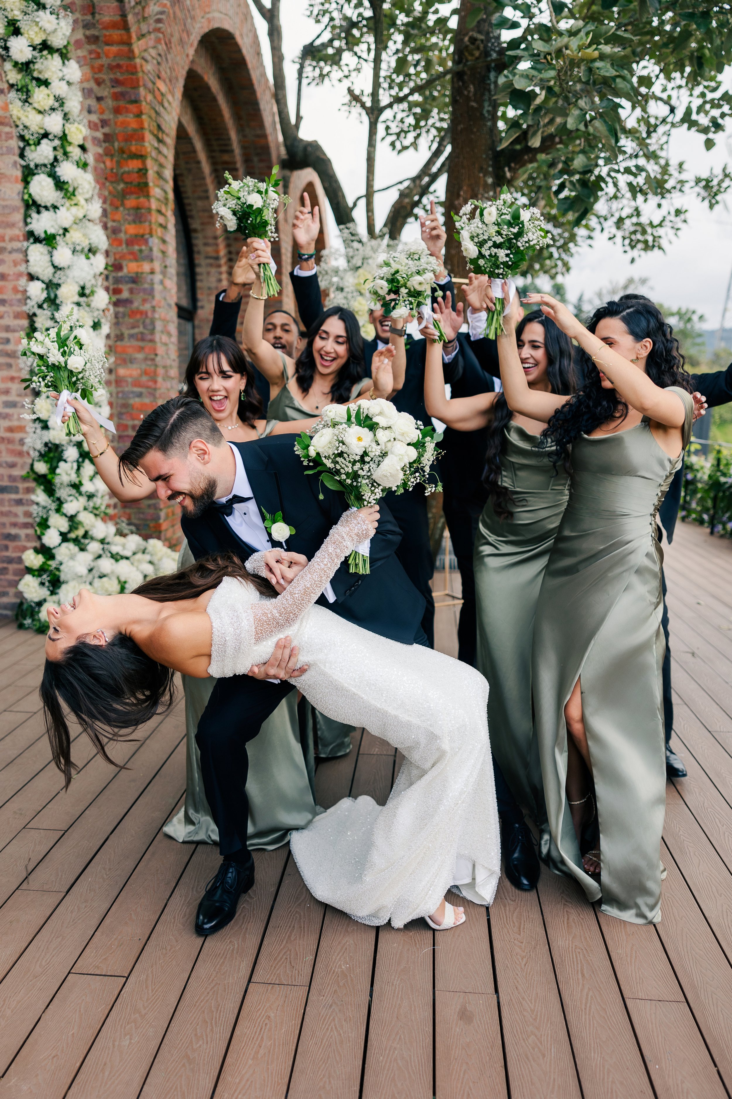 Wedding celebration with the bride and groom dancing and surrounded by bridesmaids and groomsmen, all holding bouquets, outdoors near a brick building and trees.