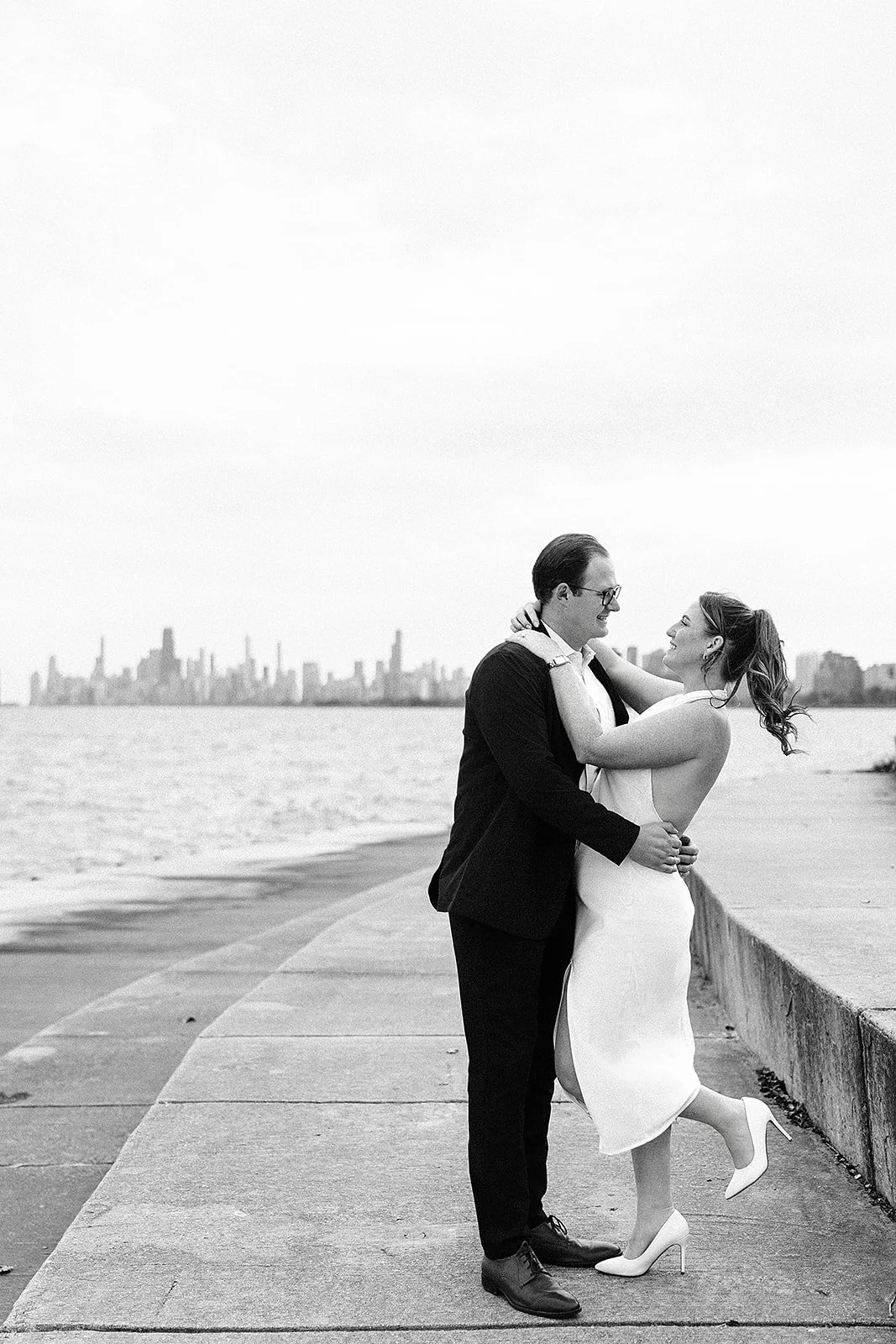 A black-and-white photo of a couple dancing by the water with a city skyline in the background.