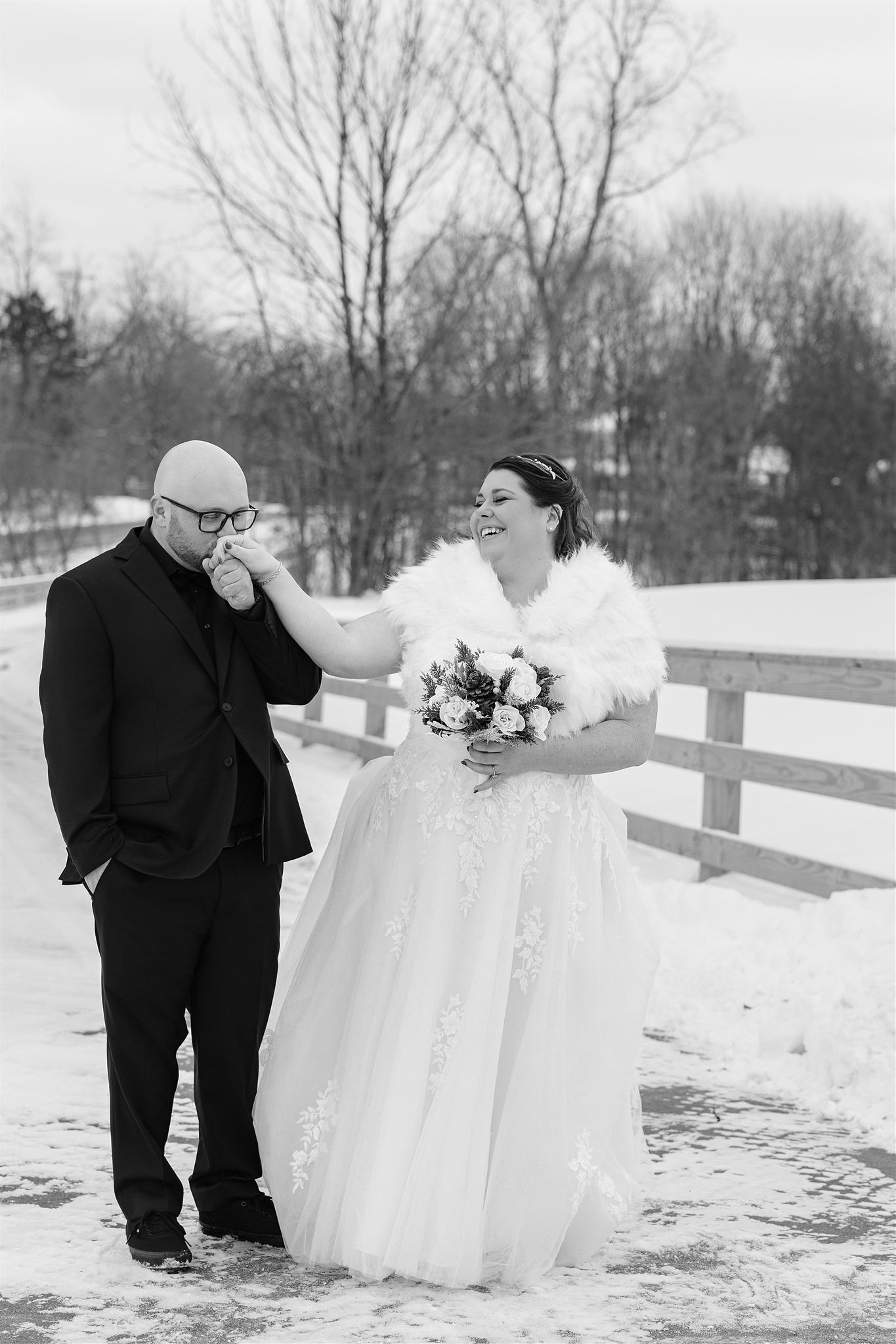 A bride in a wedding dress with a faux fur wrap holding a bouquet, laughing as she kisses a man in a suit on the forehead outside in the snow.