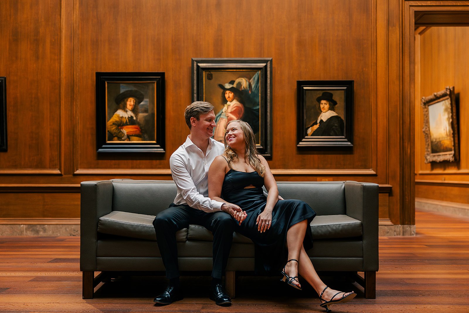 A man and woman sitting close together on a gray sofa in an art gallery, smiling and looking at each other. The gallery has wooden walls with framed classical paintings.