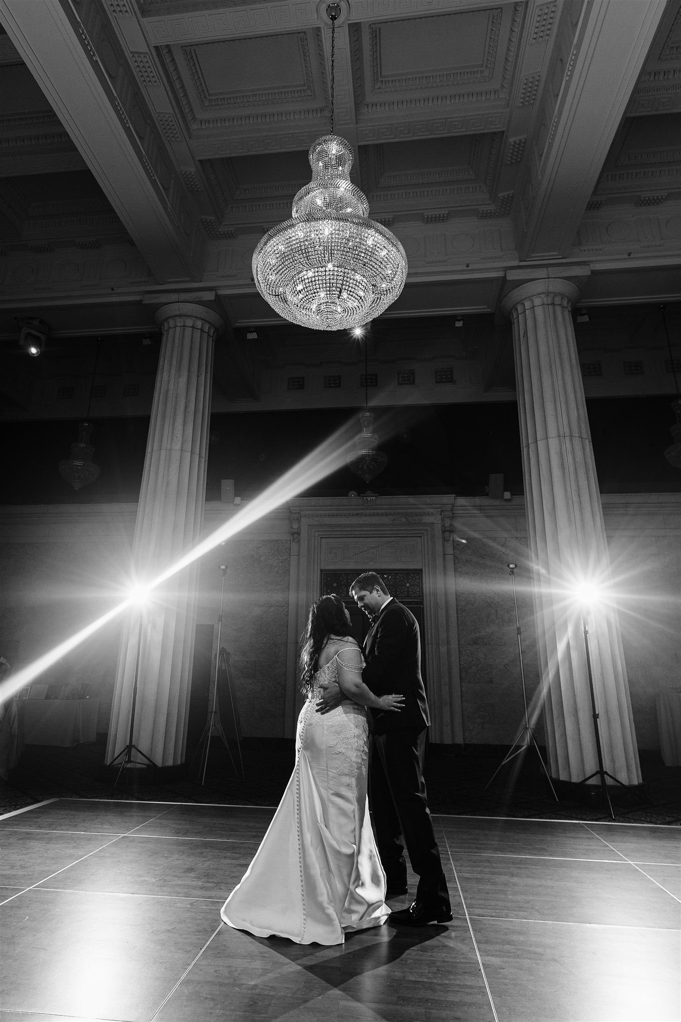 A bride and groom dancing together on a dance floor in a grand hall with large chandeliers and columns, lit by bright stage lights.