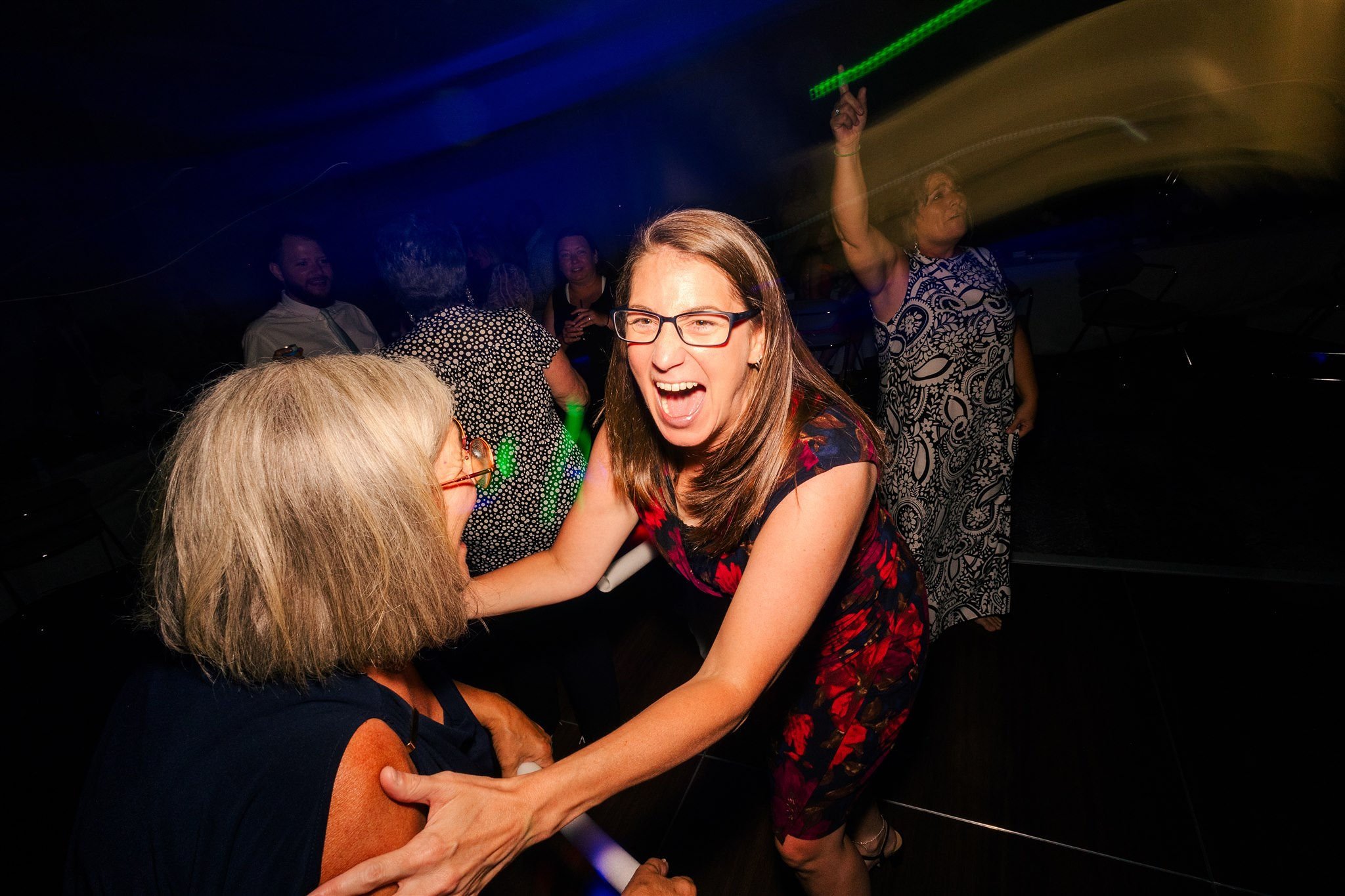Two women at a lively party, one smiling and holding the other's shoulders, with other guests dancing and laughing in the background under colorful lights.