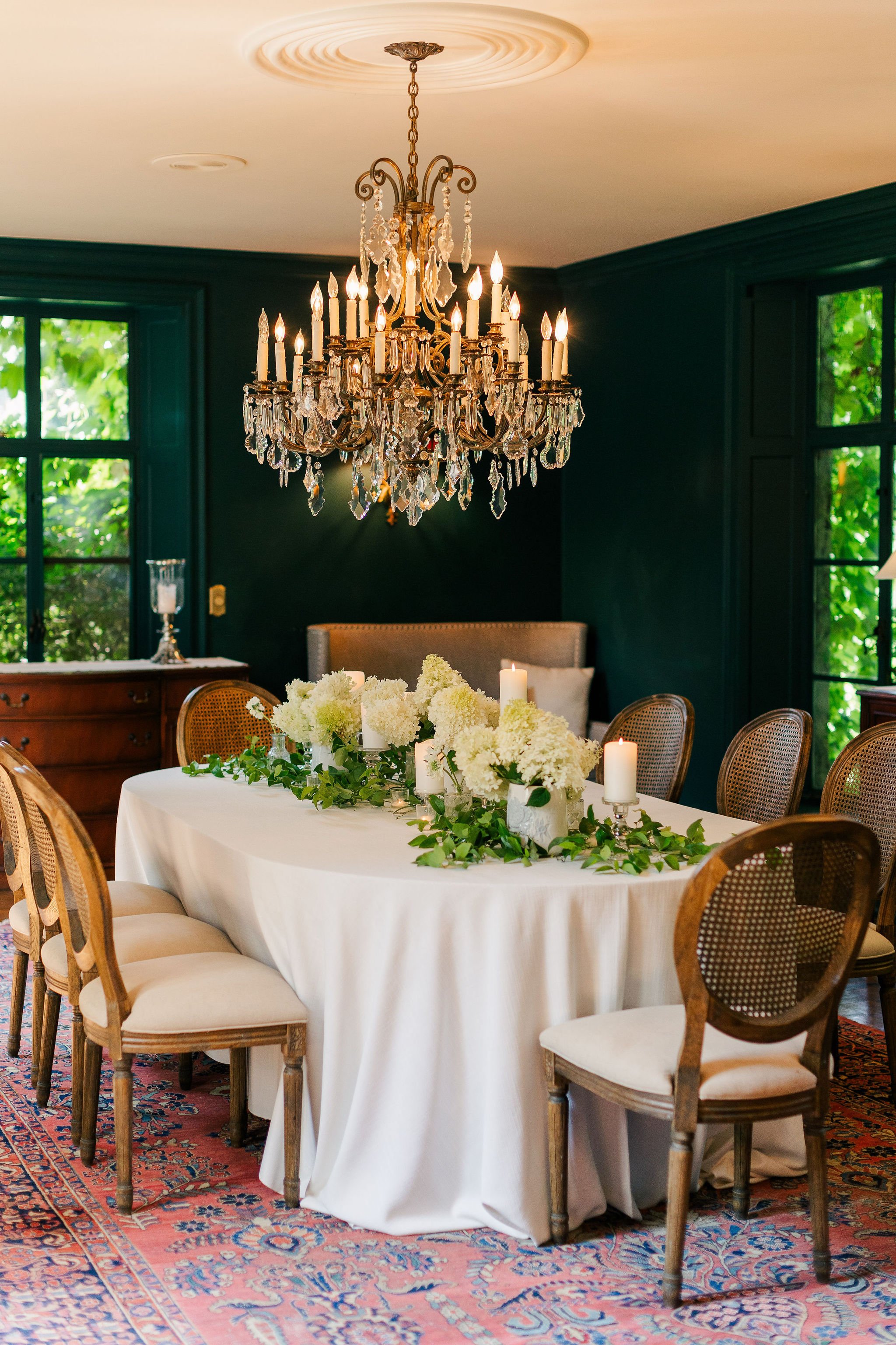 Elegant dining room with dark green walls, a large crystal chandelier, a white tablecloth-covered table with floral centerpieces, candles, and wooden chairs, with large windows showing green foliage outside.