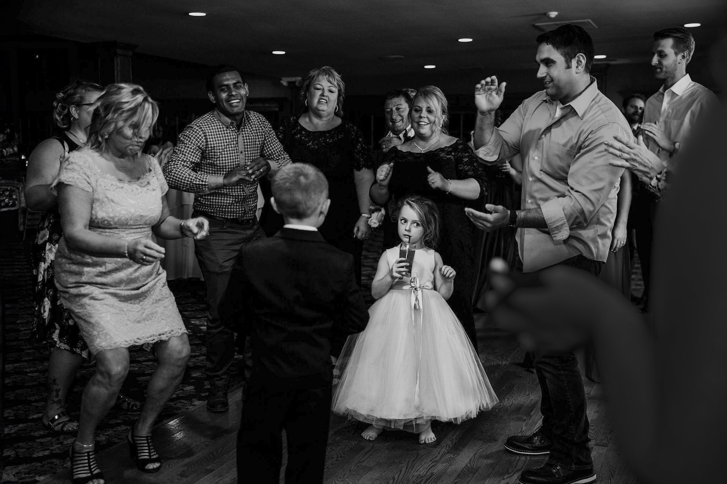 People dancing at a celebration, including a young girl in a dress drinking from a juice box, a boy in a suit, and adults enjoying the event.
