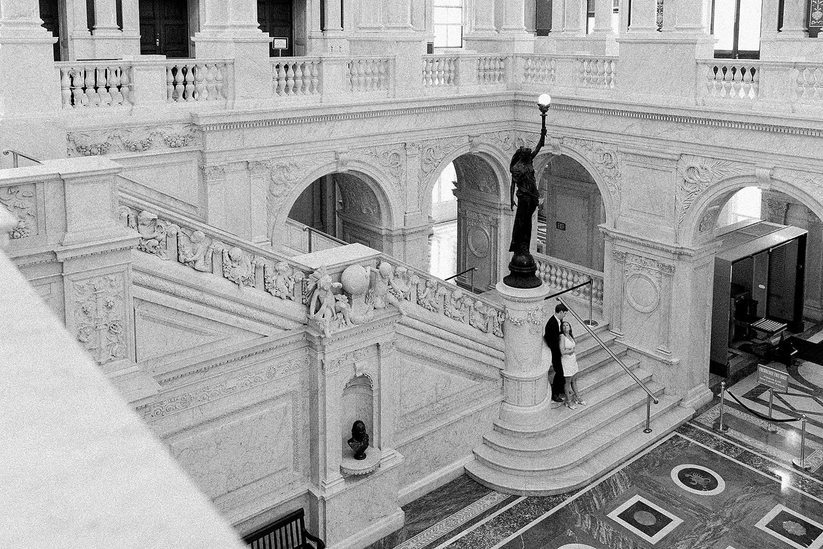 A black and white photo of an ornate interior with a marble staircase, detailed carvings, and a statue holding a lamp. Two people are standing on the stairs, leaning against the railing.