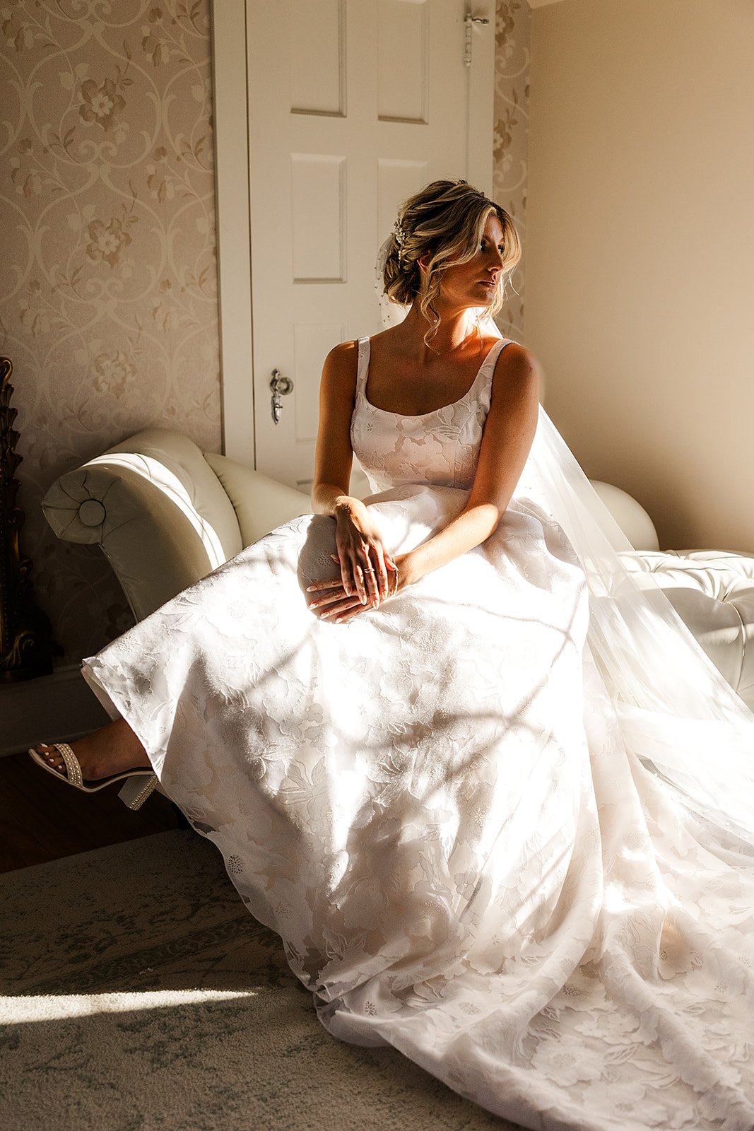 A bride in a white lace wedding dress sitting on a cream-colored vintage sofa in a sunlit room.