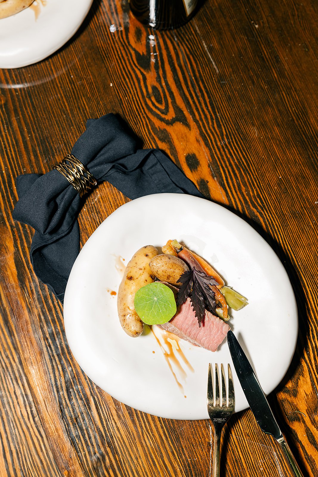 A plated dish with a piece of medium-rare meat, a roasted potato, vegetable medley, and garnishes, on a white plate on a wooden table with a black napkin and knife and fork.