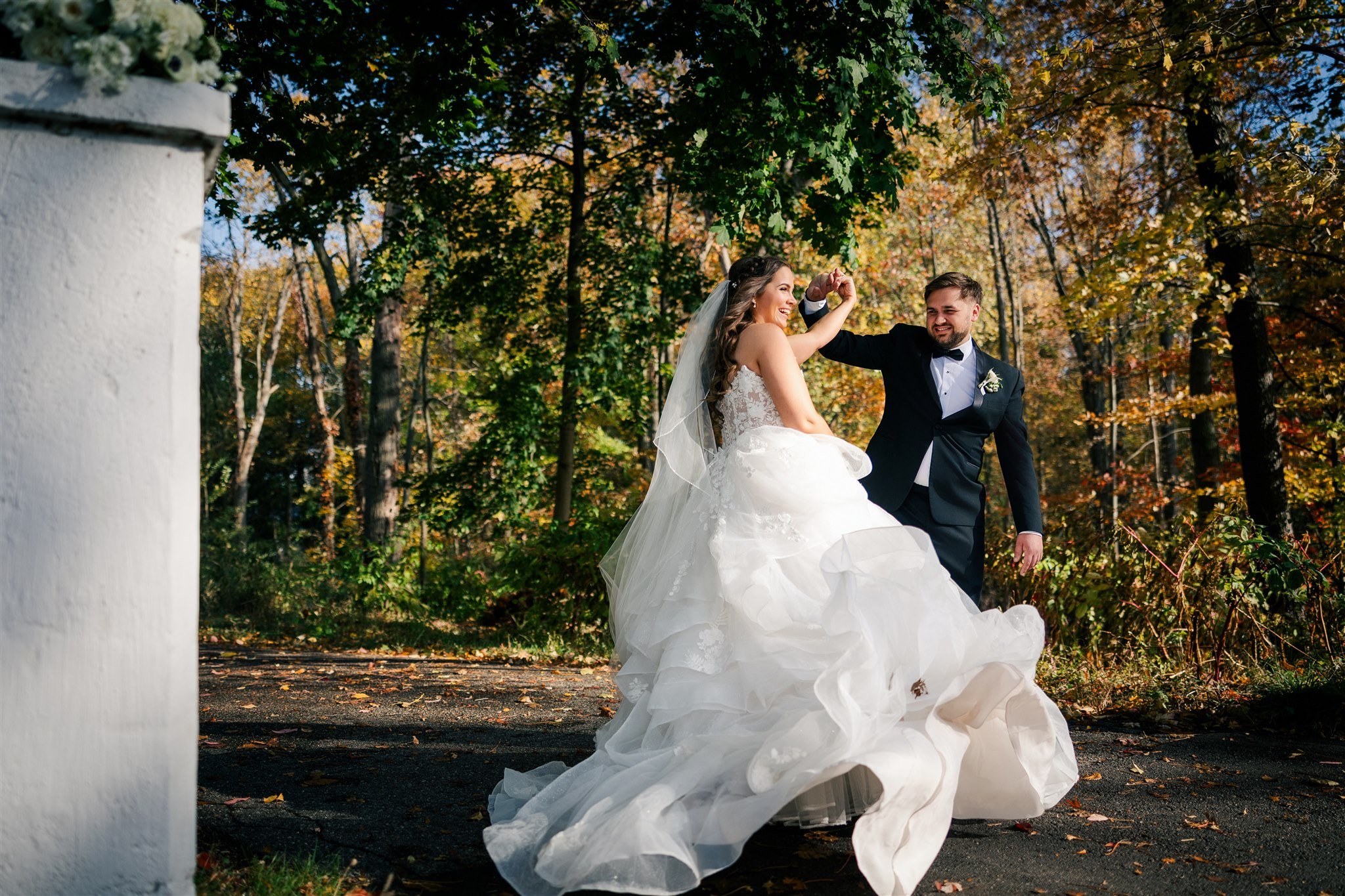 A bride in a white wedding gown and a groom in a tuxedo dancing outdoors among trees with autumn leaves, smiling and holding hands.