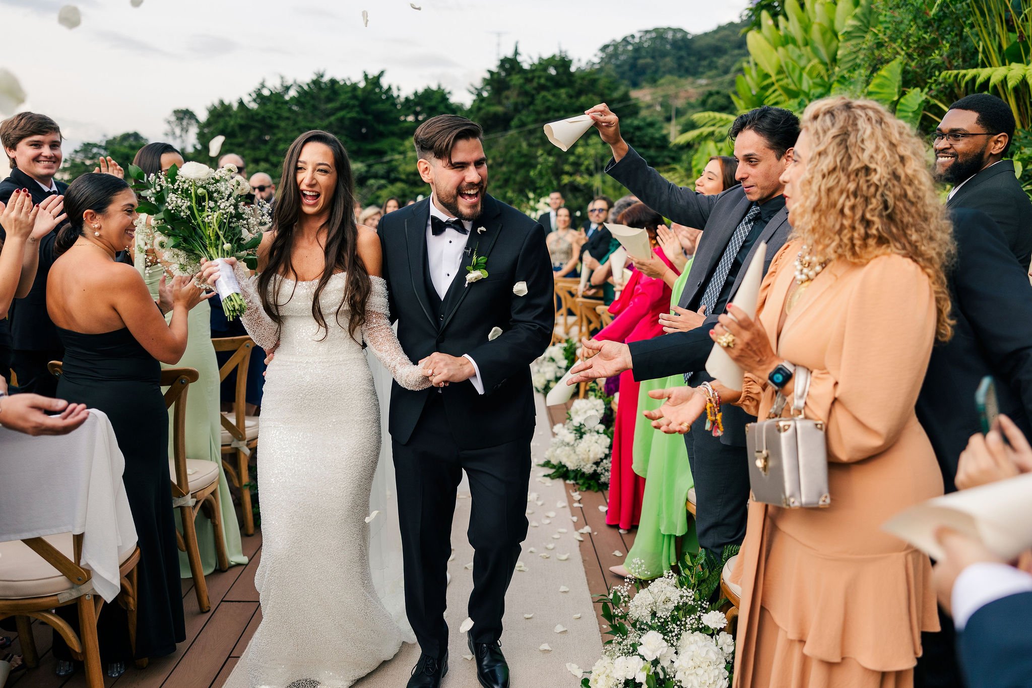 Bride and groom walking down the aisle at an outdoor wedding, surrounded by smiling guests throwing flower petals.
