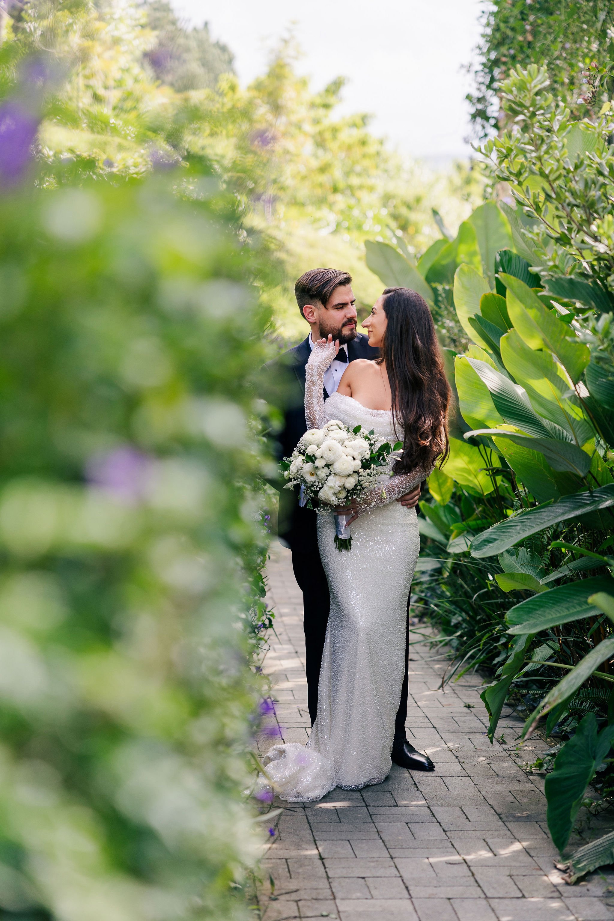 A bride and groom standing close together on a garden pathway, surrounded by lush greenery, sharing an intimate moment on their wedding day.