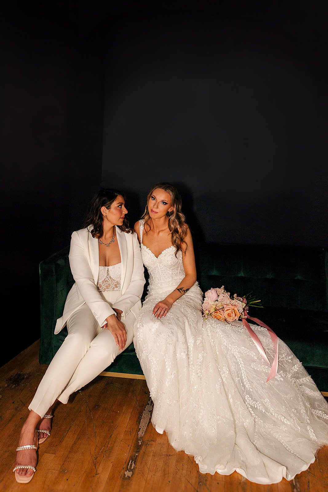 Two women sitting on a green velvet bench, one in a white suit and the other in a white wedding dress, with a bouquet of pink roses beside them, against a black wall backdrop.