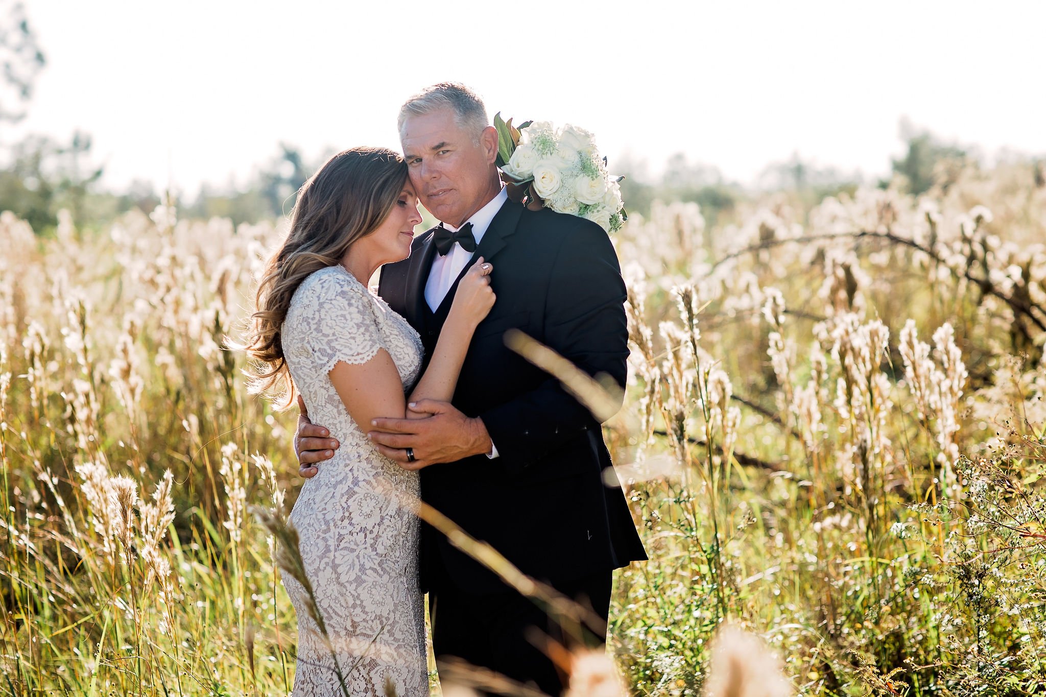 A bride and groom embrace in a field of tall grasses during sunset, with the bride resting her head on the groom's chest and both holding each other's arms.